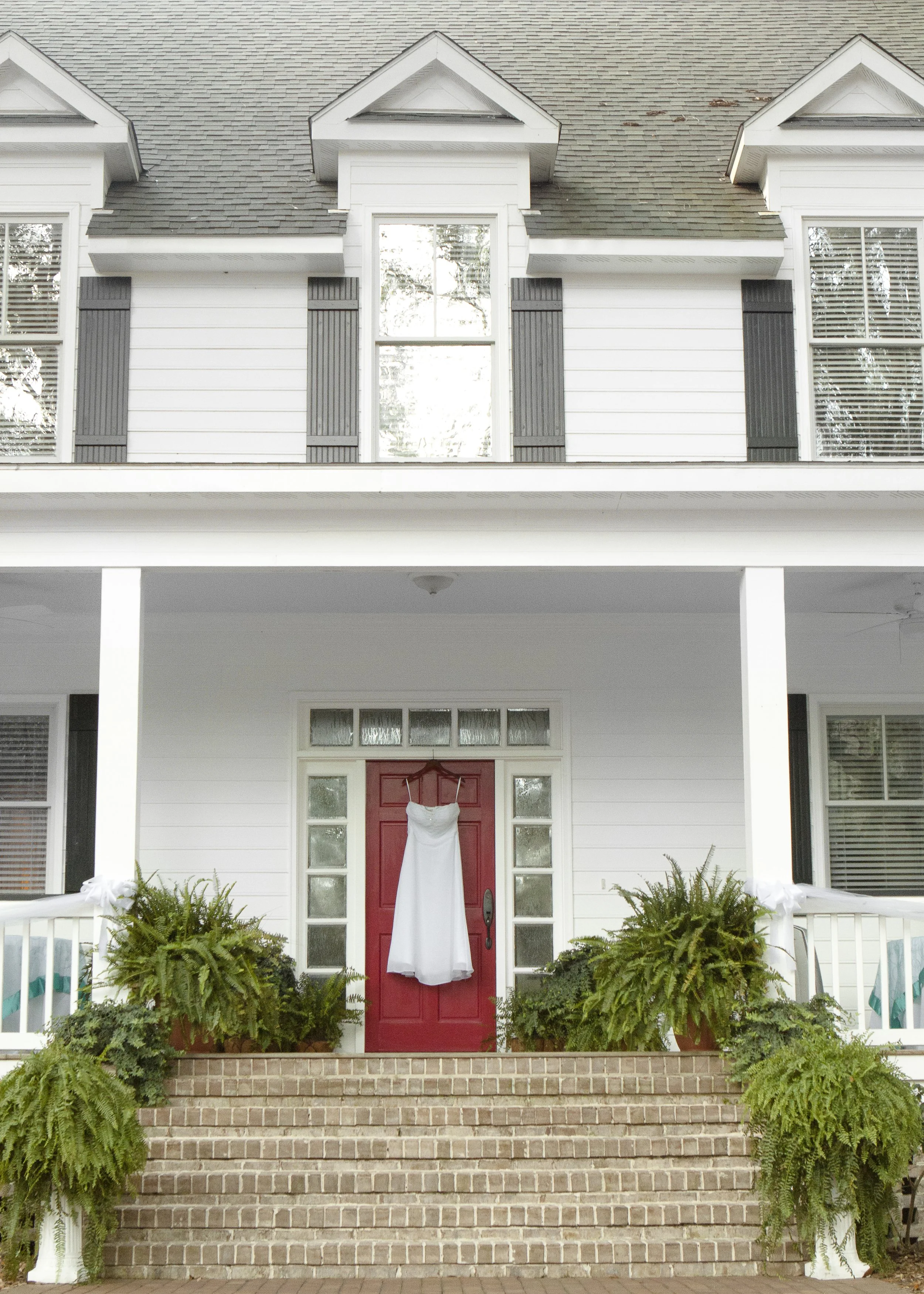 A white wedding dress hanging on a hook on a red front door of a house with brick steps leading up to it. Green plants are on either side of the steps.