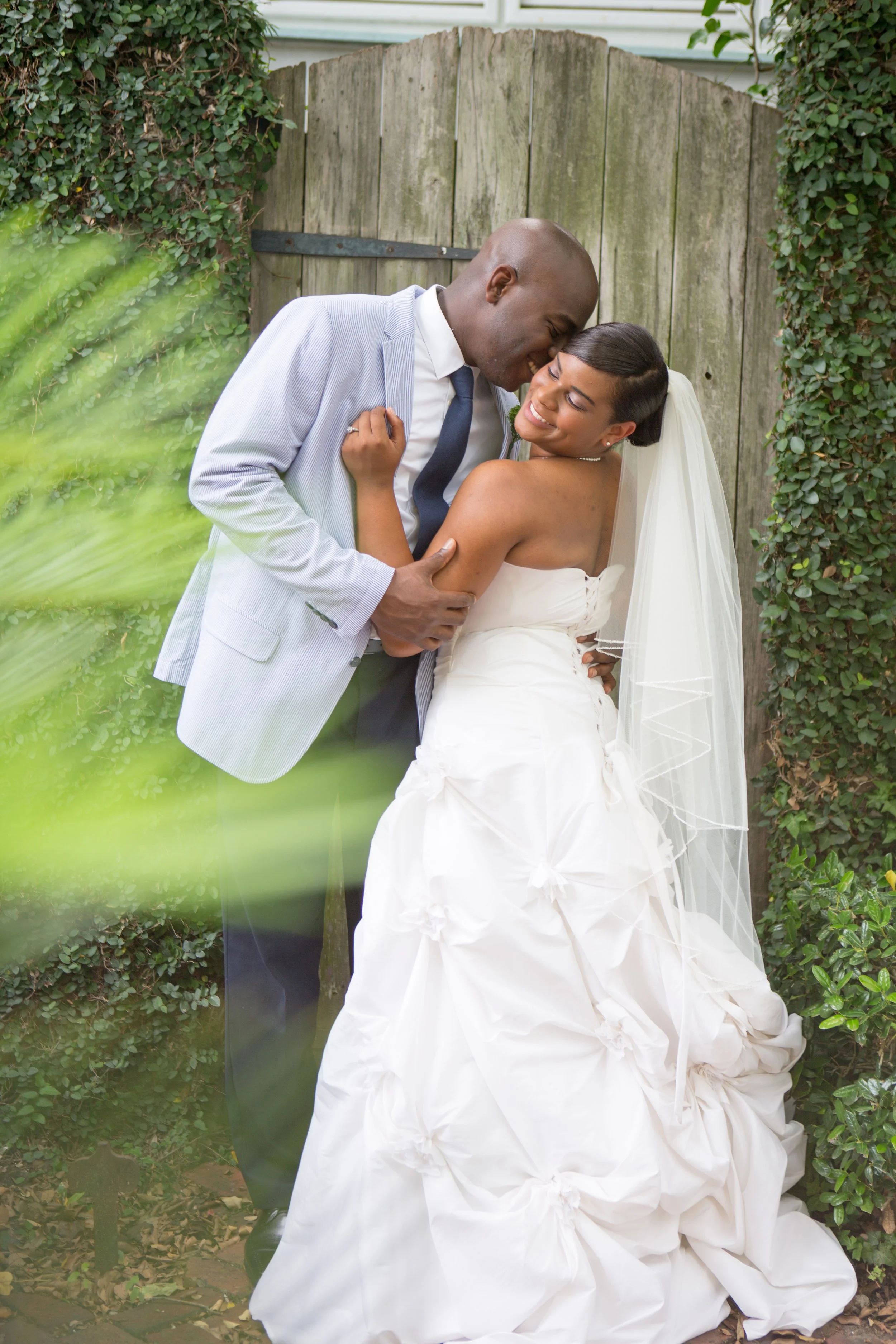 A bride and groom sharing a joyful, intimate moment outdoors in front of a wooden fence, surrounded by greenery.