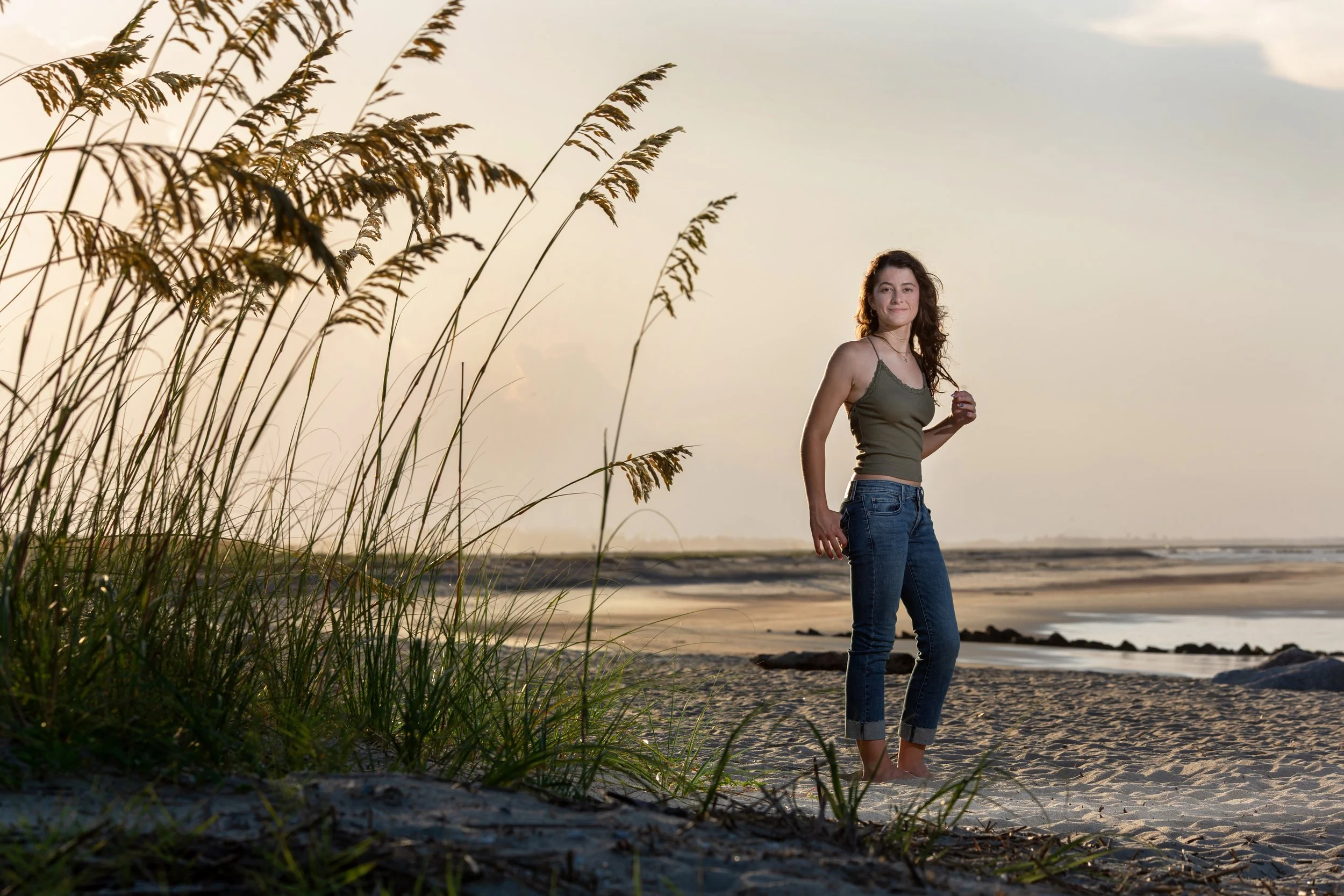 A young woman with curly hair stands barefoot on a sandy beach during sunset, wearing a green tank top and rolled-up jeans, with tall grass in the foreground.