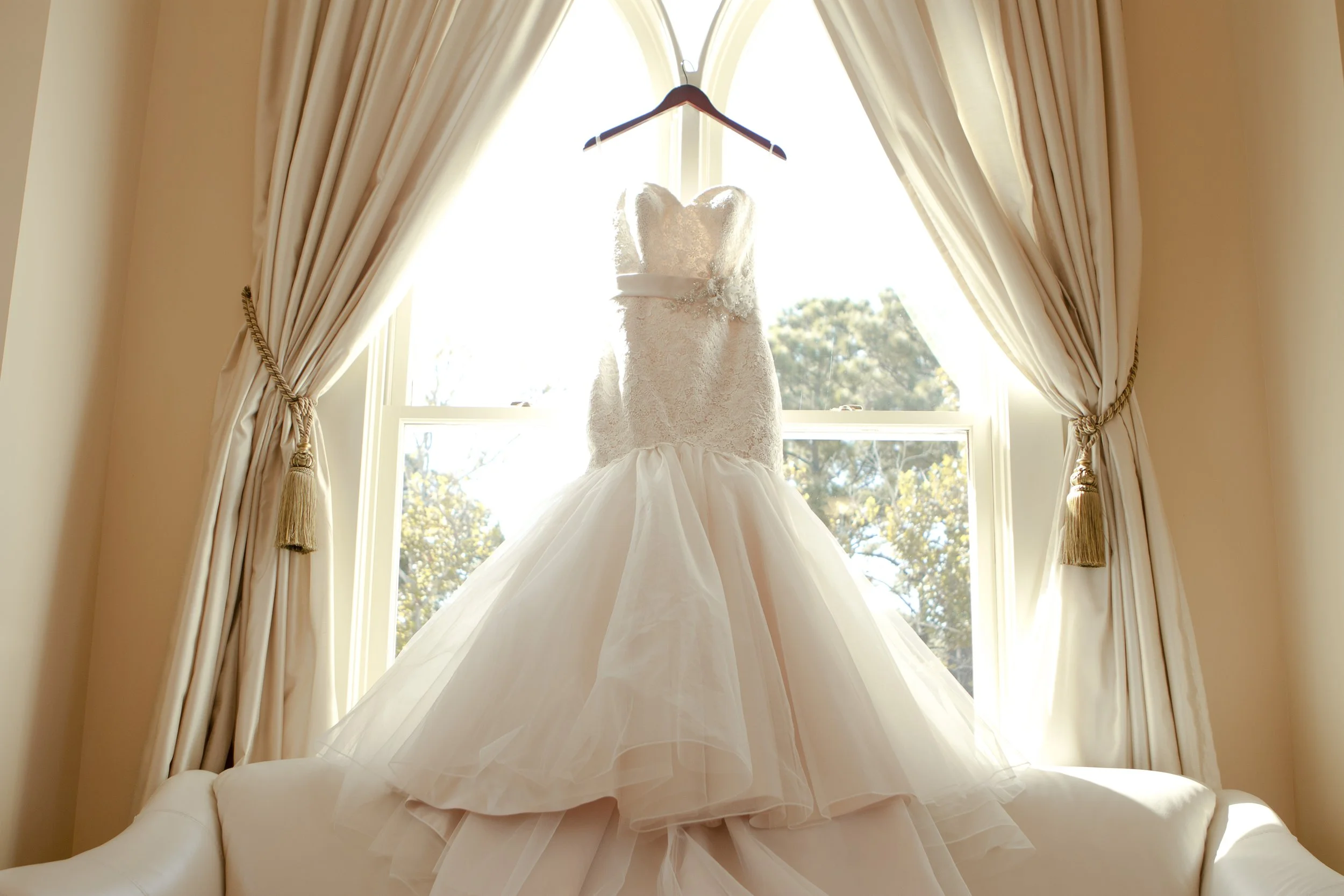A white wedding dress with lace bodice and full tulle skirt hanging in front of a window with cream-colored curtains, illuminated by natural sunlight.