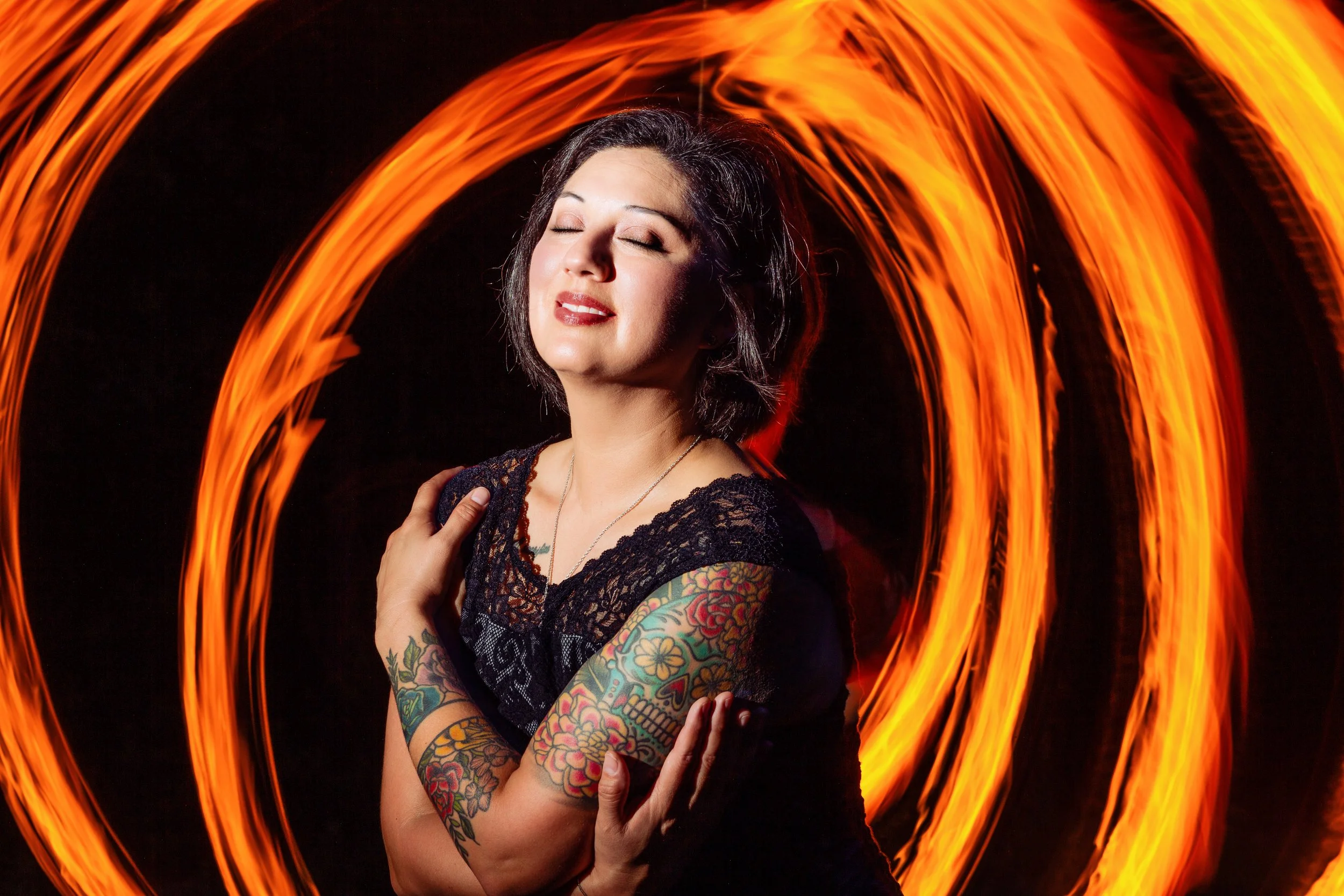 Woman with dark hair, tattoos on arms, wearing a black lace top, smiling with eyes closed, surrounded by swirling orange light trails