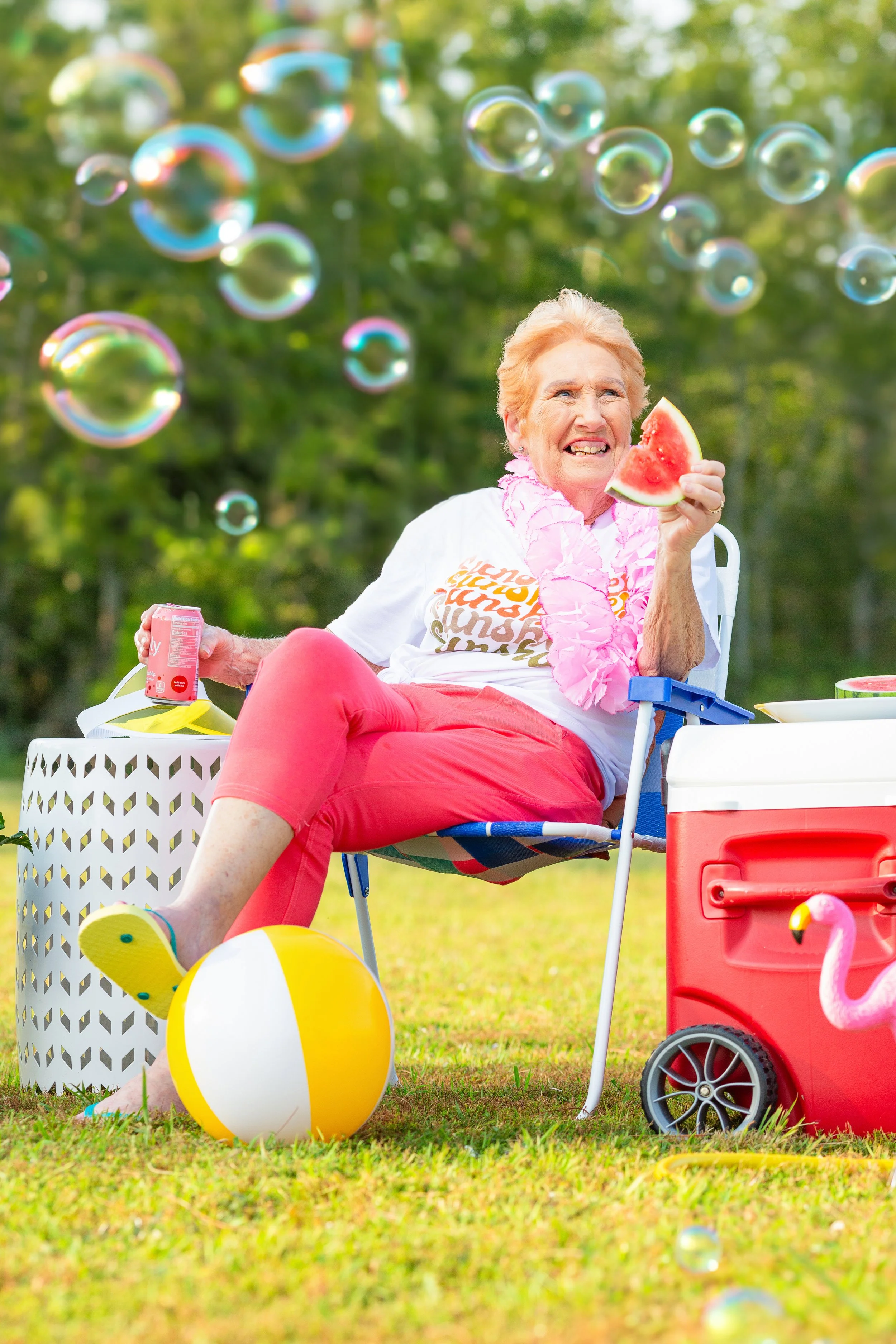 A joyful elderly woman sitting in a chair outdoors. She is holding a slice of watermelon, surrounded by soap bubbles and summer picnic items like a beach ball, a cooler, and a can of soda.