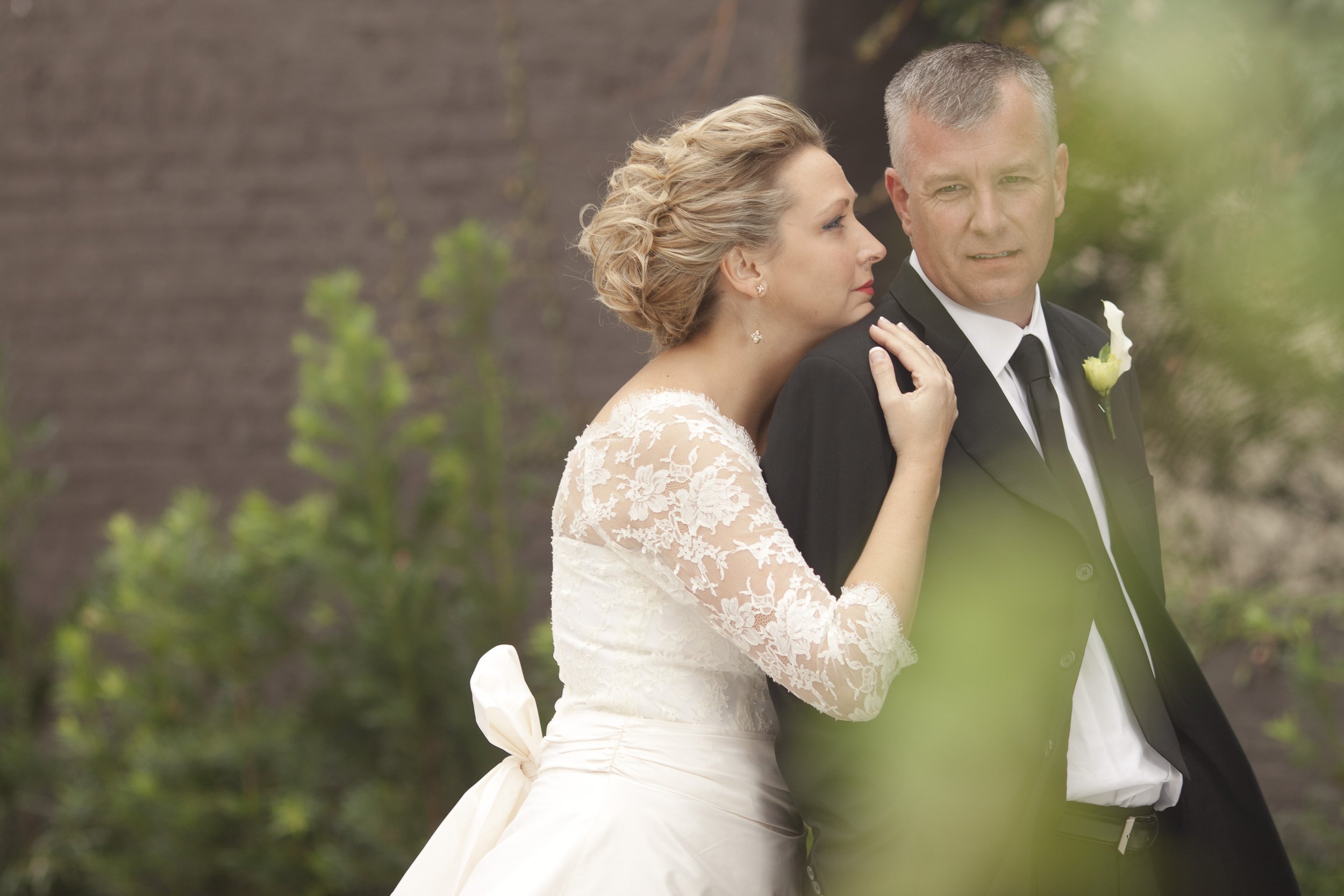 A bride in a wedding dress and a groom in a tuxedo stand close together outdoors, with the bride resting her head on the groom's shoulder.