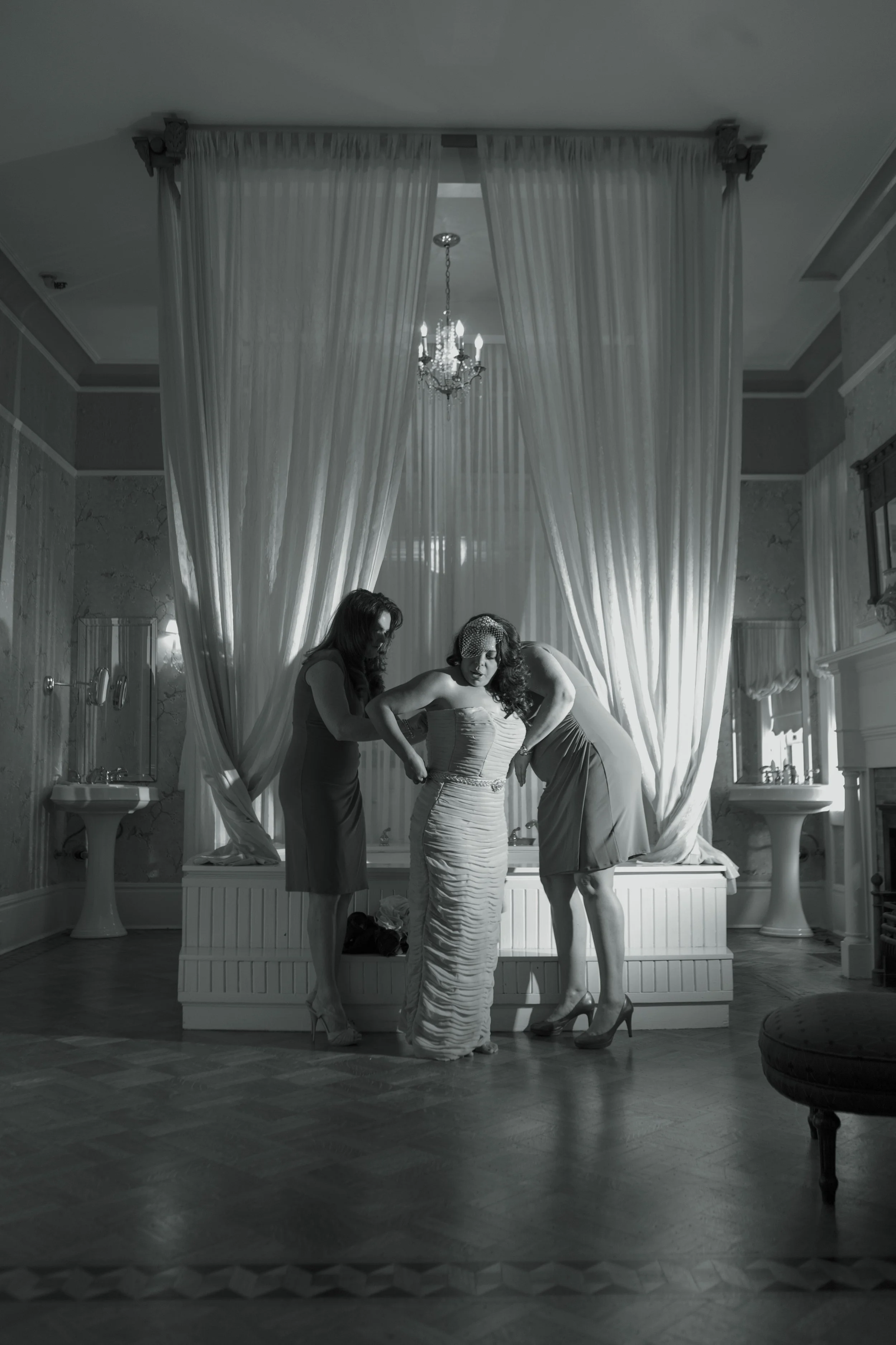 A bride trying on a wedding dress with two women assisting her in a luxurious room with elegant drapery, chandeliers, and vintage decor.