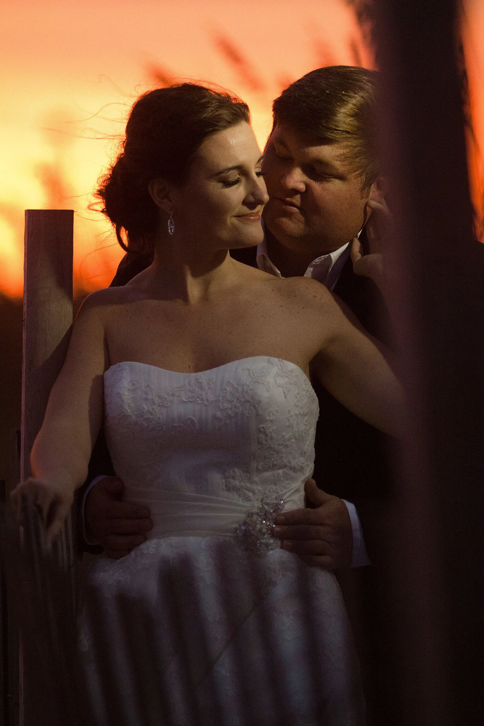 A woman in a white wedding dress and a man in a dark suit holding each other, sharing an intimate moment during sunset.