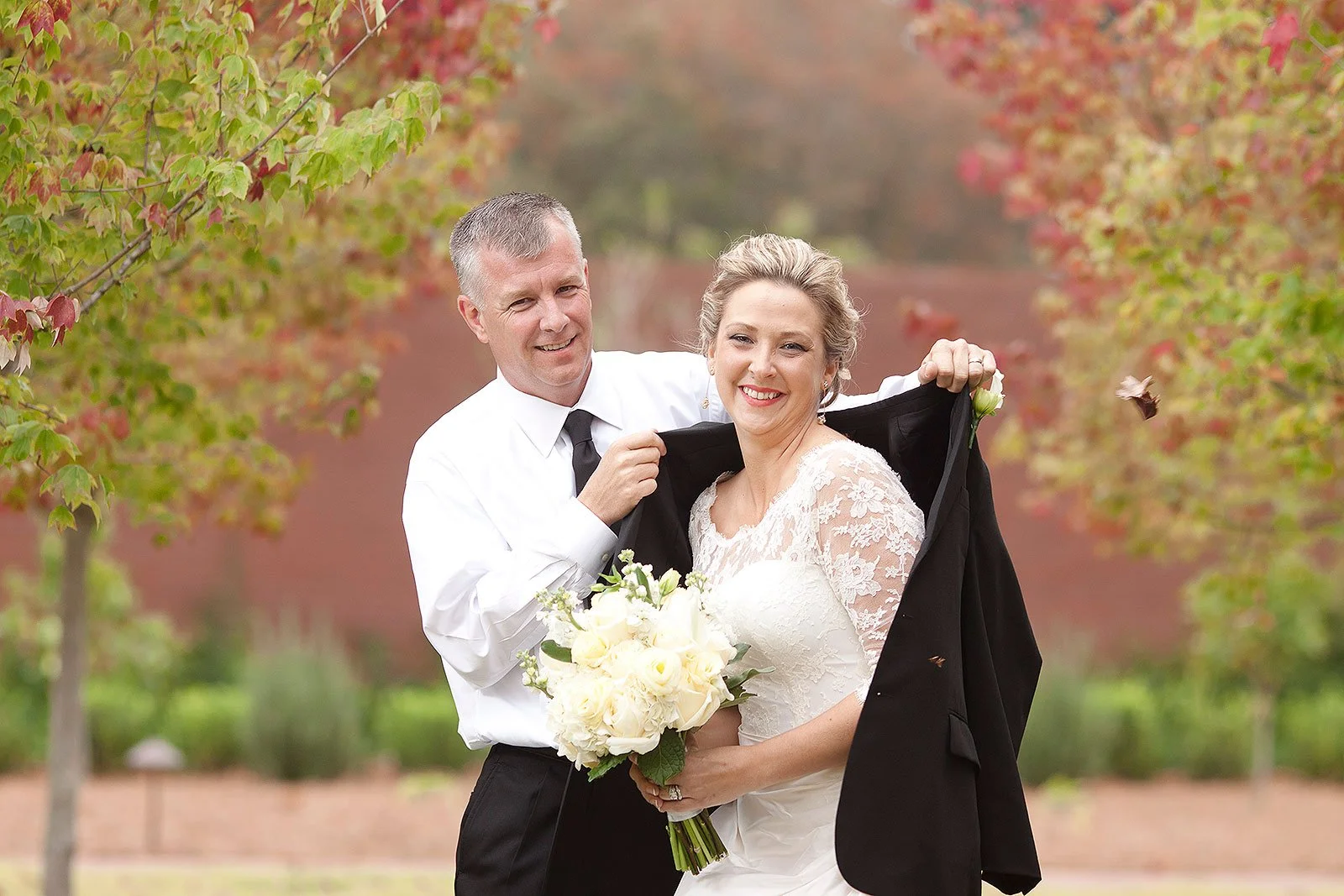 A joyful couple in wedding attire outdoors, with the groom holding a suit jacket over the bride's shoulders, and the bride holding a bouquet of white flowers, surrounded by trees with colorful autumn leaves.