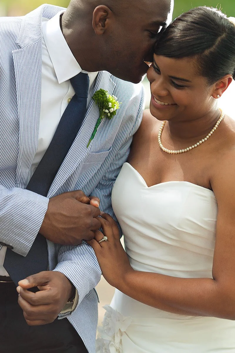 A happy couple at their wedding, with the groom gently kissing the bride's forehead. The bride is smiling, wearing a white dress and a pearl necklace, while the groom is dressed in a light blue striped suit with a white shirt and navy tie, and a bout