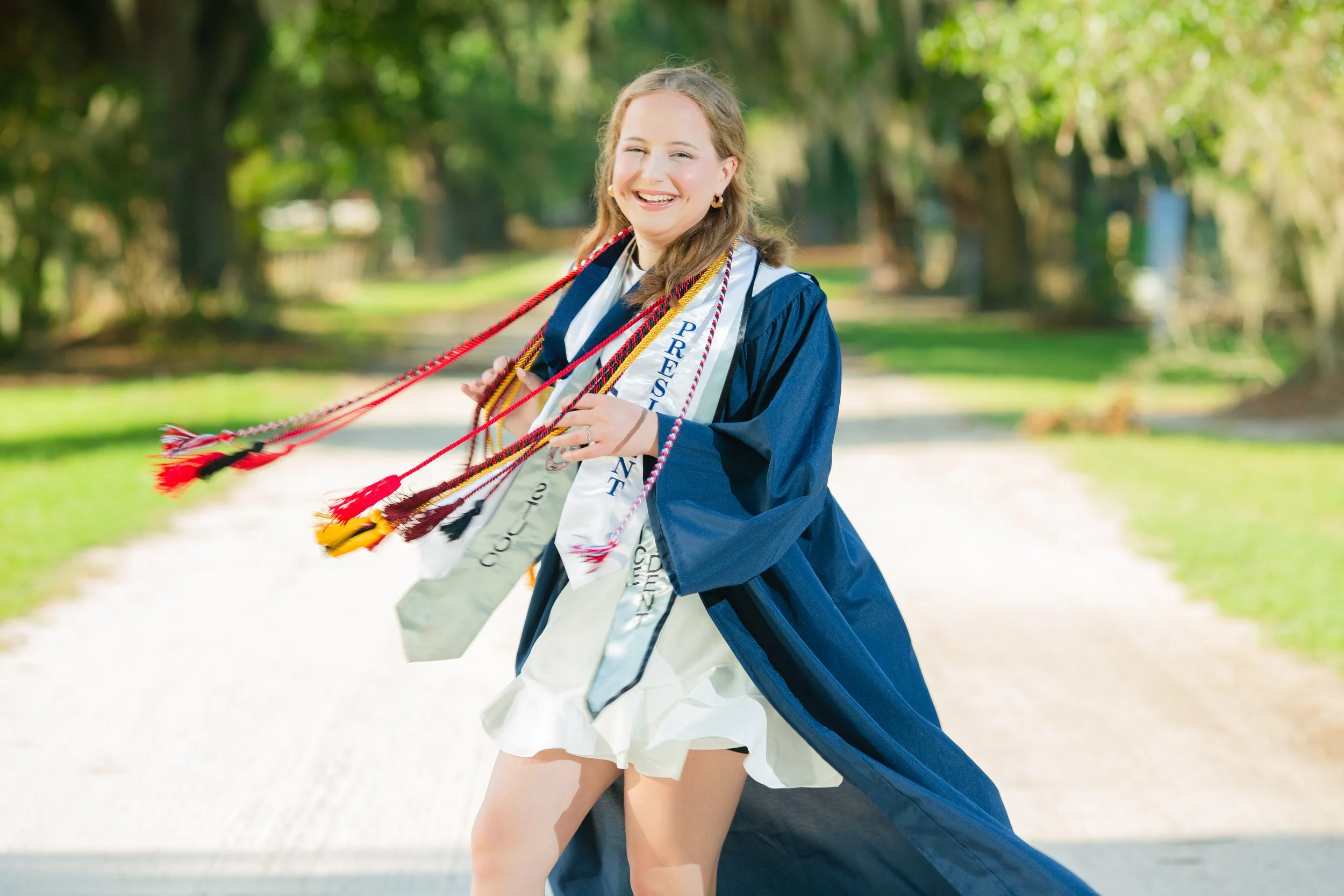 A young woman in a graduation gown and cap, celebrating on a pathway with trees in the background, wearing honor cords and sashes.