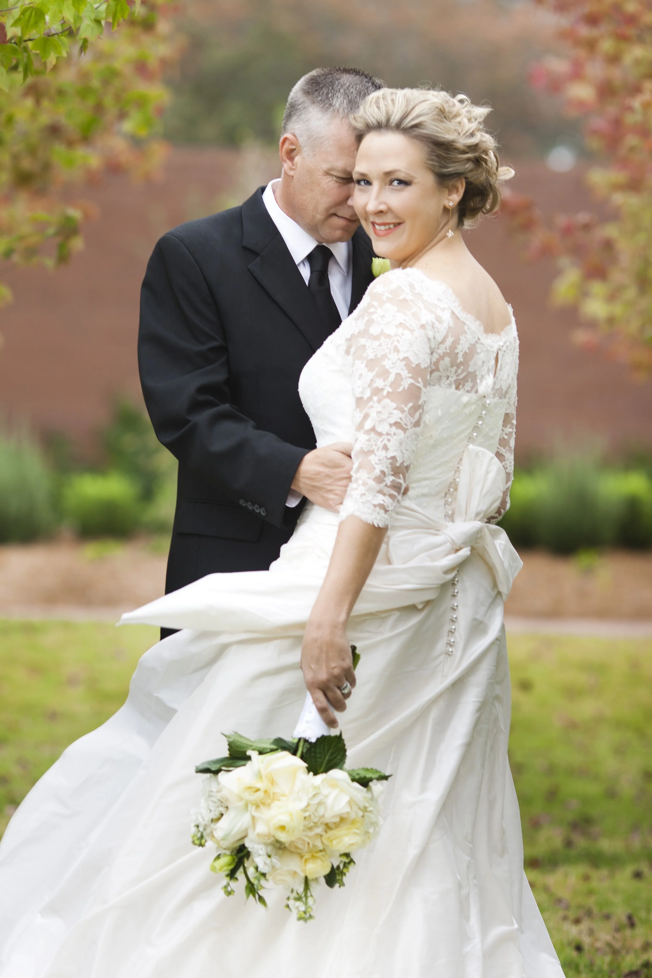 A bride in a white wedding dress holding a bouquet of white roses, standing close to a man in a black tuxedo, with the man tenderly holding the bride.