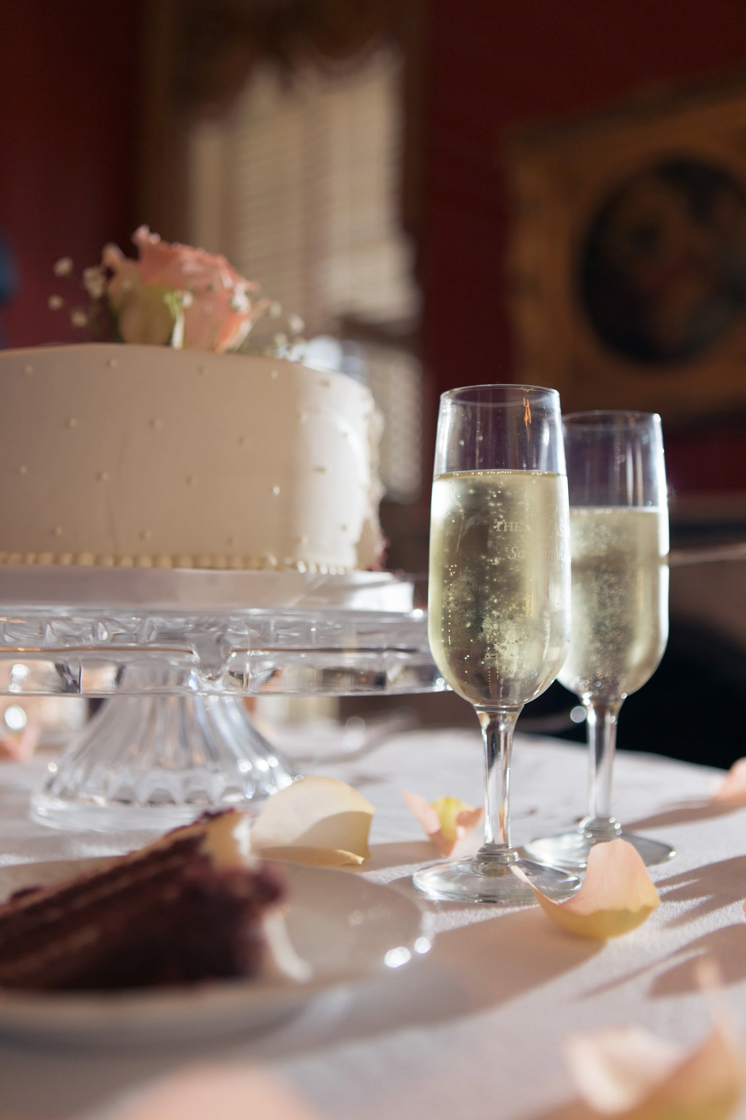 A wedding celebration scene featuring a white cake with pink roses, two glasses of sparkling wine, a slice of chocolate cake, and scattered rose petals on a table.
