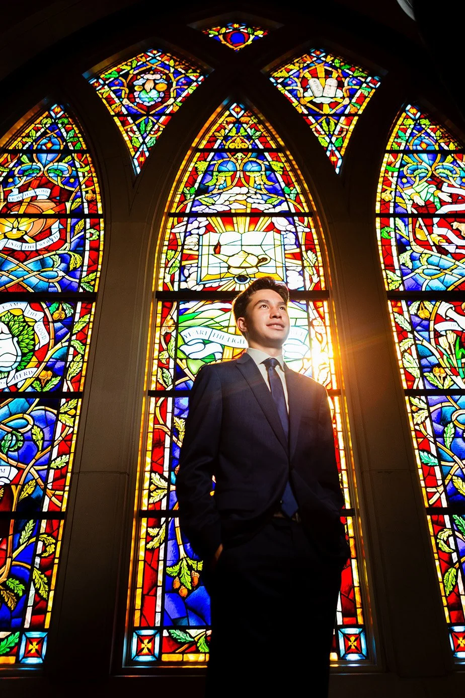 Young man in a dark suit standing in front of colorful stained glass windows with sunlight shining through.