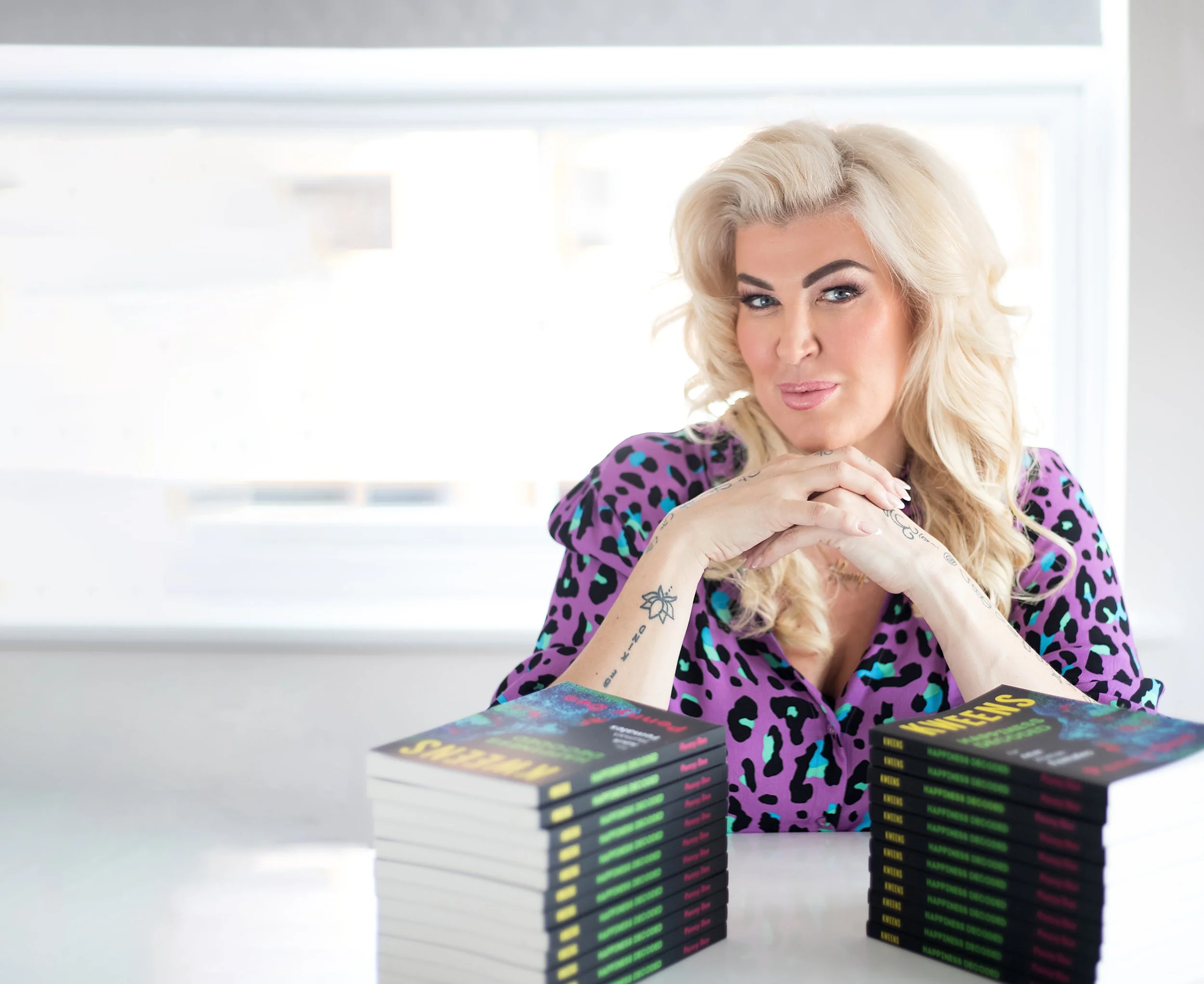 A woman with blonde, curly hair and tattoos on her arms, wearing a purple leopard print top, sitting at a white table with stacks of books with colorful, neon-style covers in front of her, near a window.