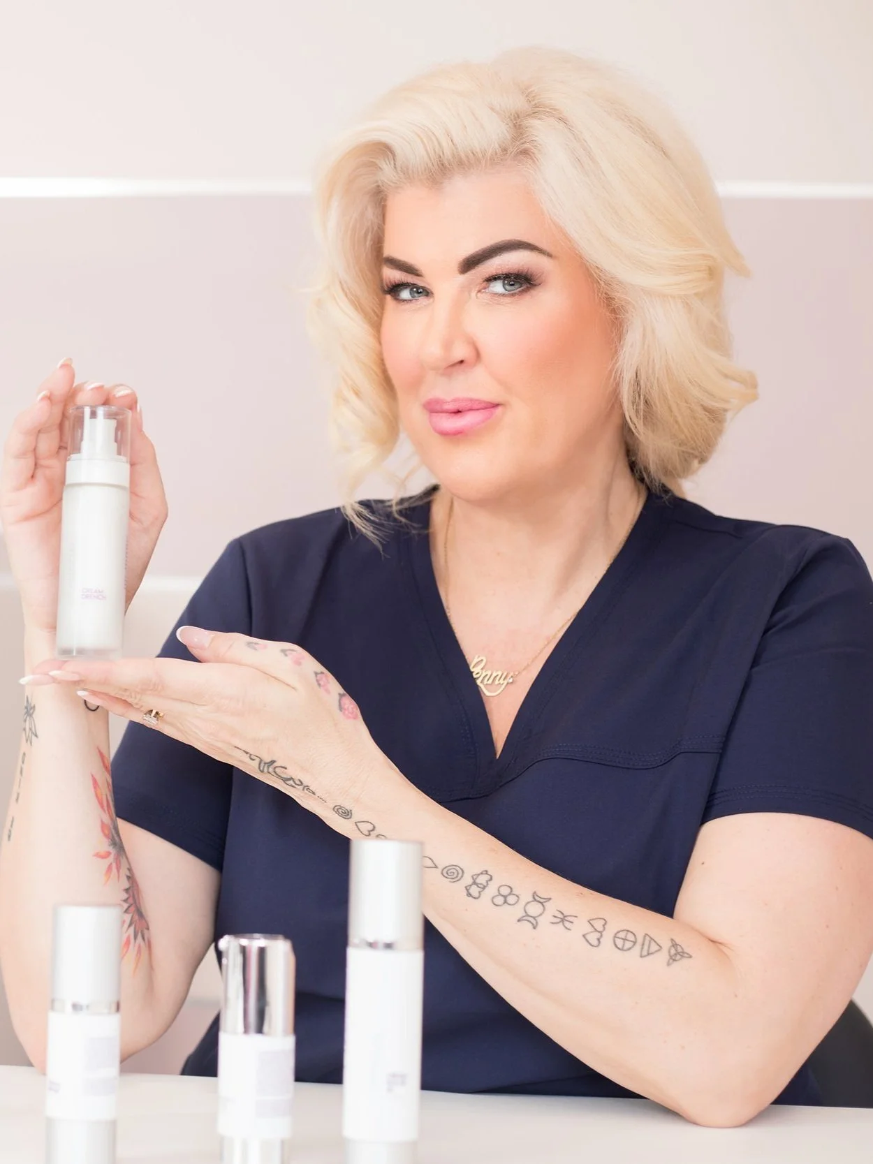 A woman with platinum blonde hair and tattoos on her arms, wearing navy scrubs, holding a skincare product in a white bottle with a clear cap, sitting at a table with multiple similar skincare bottles, and smiling at the camera.