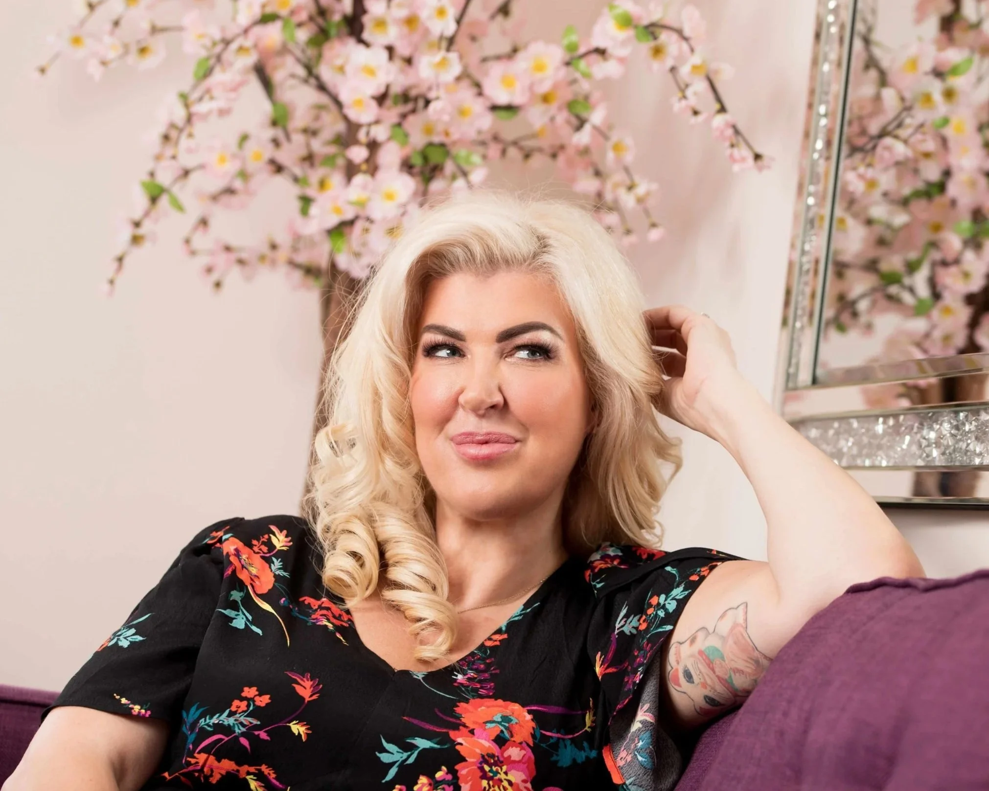 A woman with blonde, curled hair sitting on a purple couch, smiling thoughtfully. She is wearing a black floral top and has a tattoo on her arm. Behind her, there is a pink wall with a large branch of pink cherry blossoms and a mirror with a glittery frame.