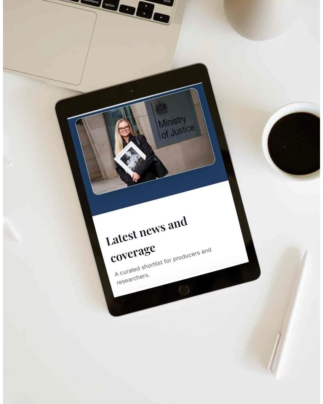 A tablet displaying a webpage with a woman standing outside a building labeled 'Ministry of Justice', holding a newspaper. The webpage has a section titled 'Latest news and coverage'. The tablet is on a white desk with a cup of coffee, a silver laptop, and a white pen nearby.