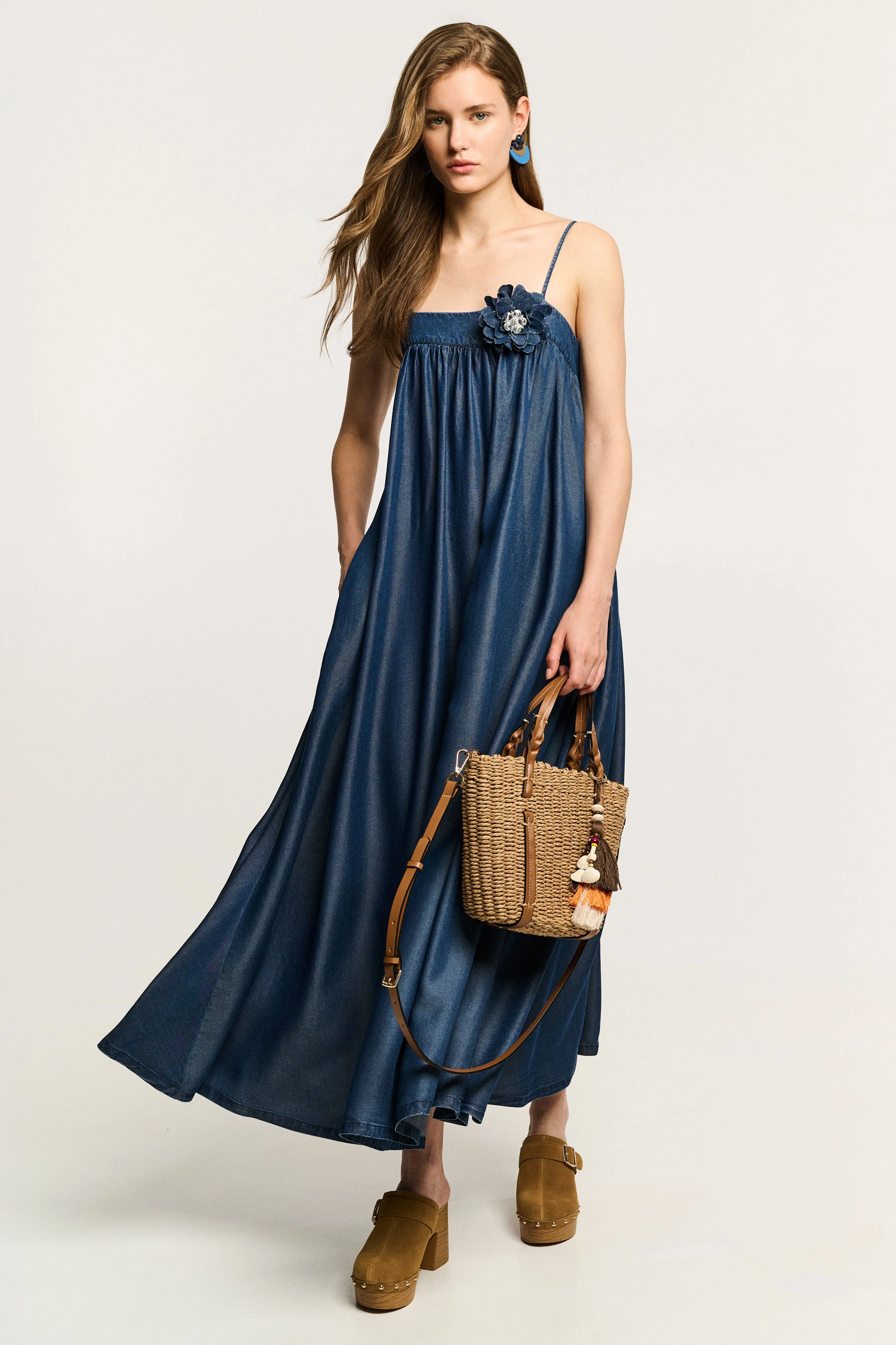 A young woman with long hair wearing a blue maxi dress with spaghetti straps and floral embellishment, carrying a woven handbag, and wearing tan platform heels, stands against a plain background.