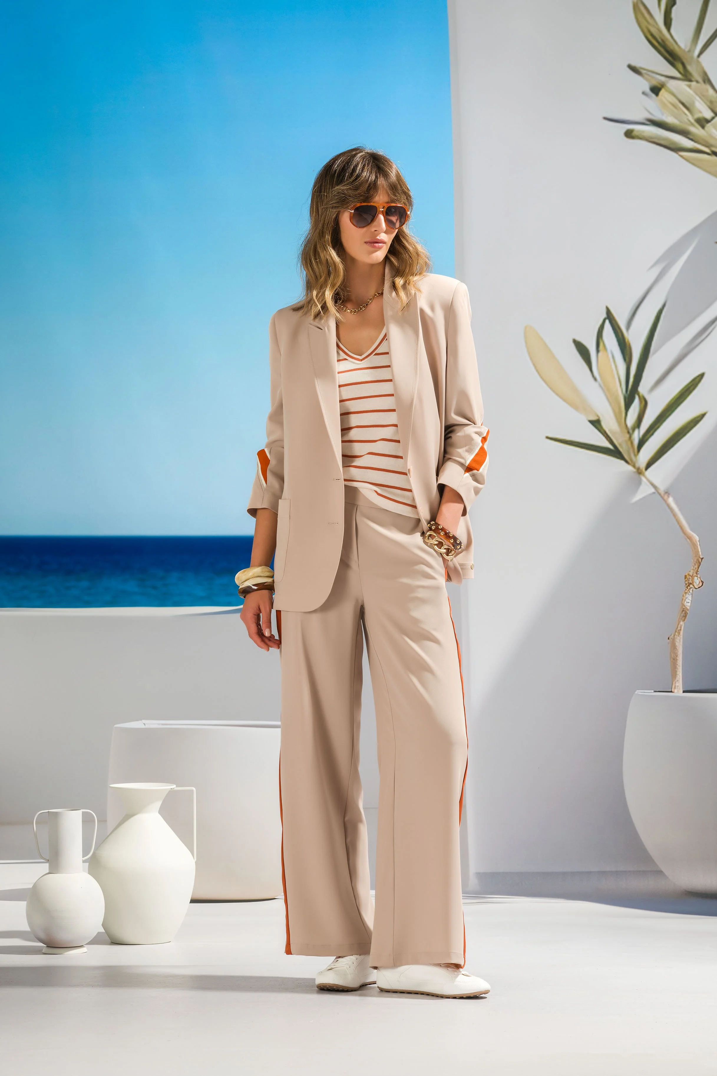 A woman wearing beige pants, a beige blazer, a striped shirt, white shoes, large sunglasses, and jewelry, standing in a bright minimalist setting with white vases, a plant, and a backdrop of a blue sky and ocean.