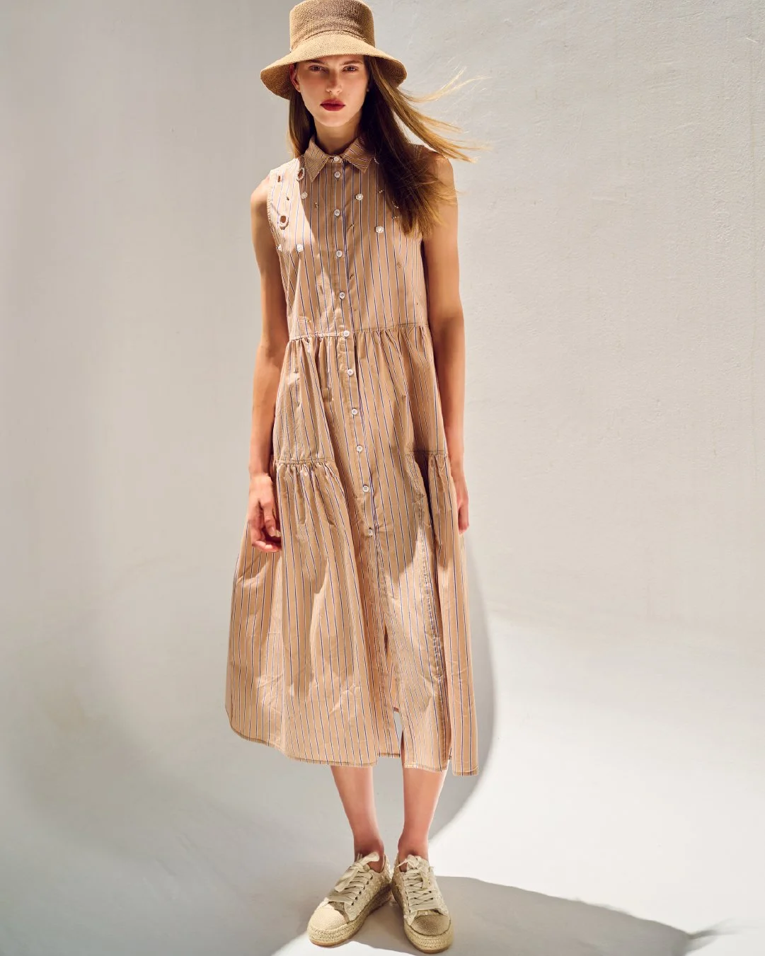 Young woman in a beige and white striped sleeveless dress, wearing a beige hat and sneakers, standing against a plain light-colored background.