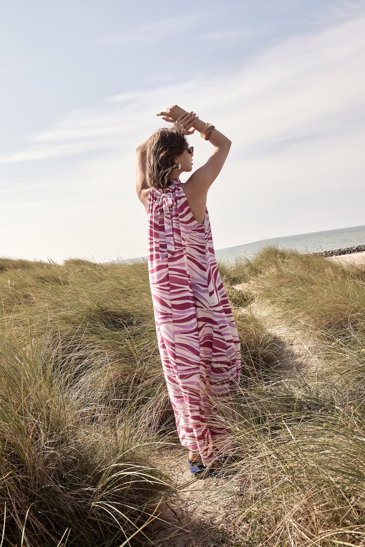 A woman in a patterned pink and white dress, wearing sunglasses and jewelry, standing in a grassy coastal landscape with the ocean and sky in the background, stretching her arms upward.