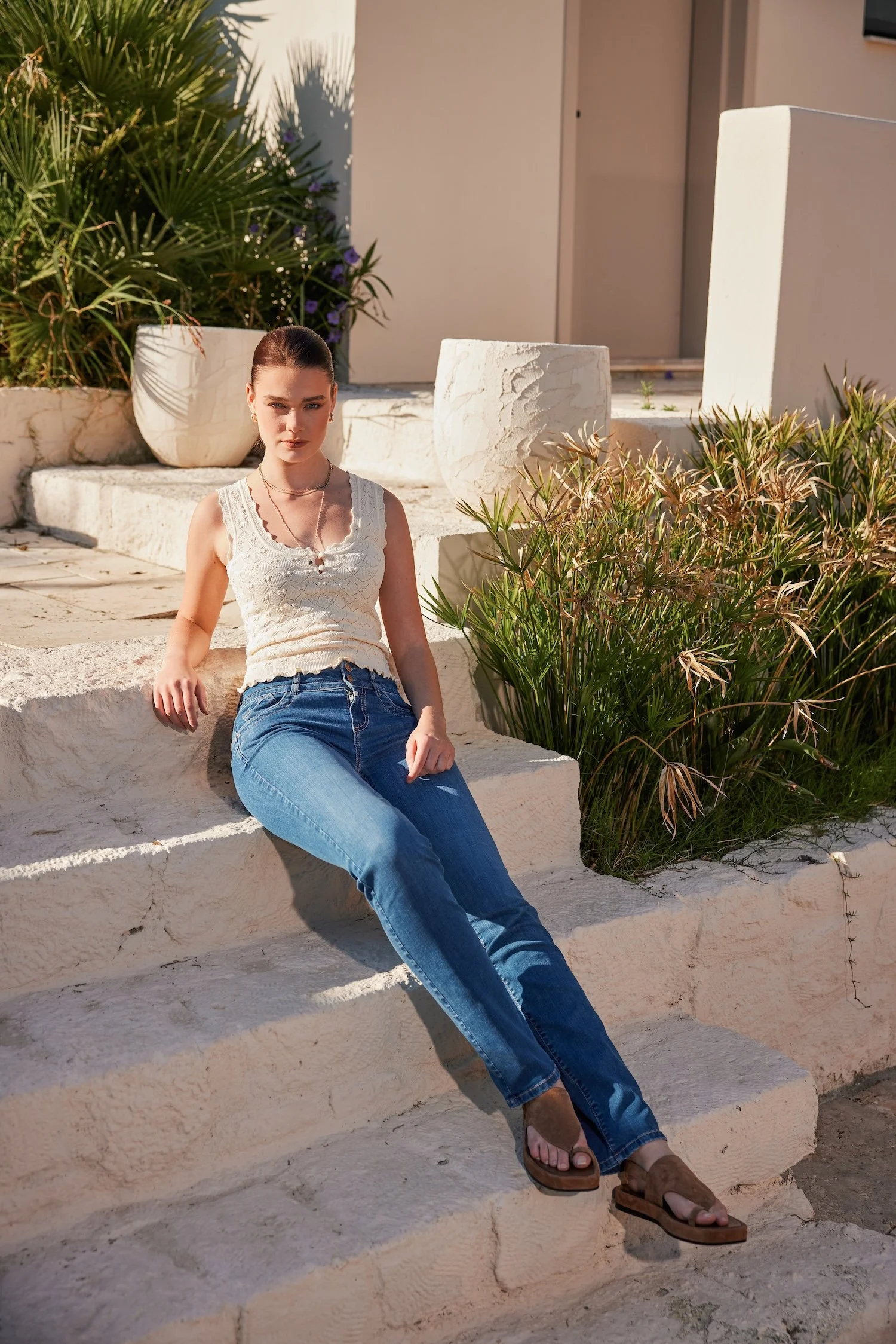 A young woman with brown hair tied in a bun, wearing a white sleeveless top, blue jeans, and brown sandals, sitting on stone stairs outside a beige building with greenery around.