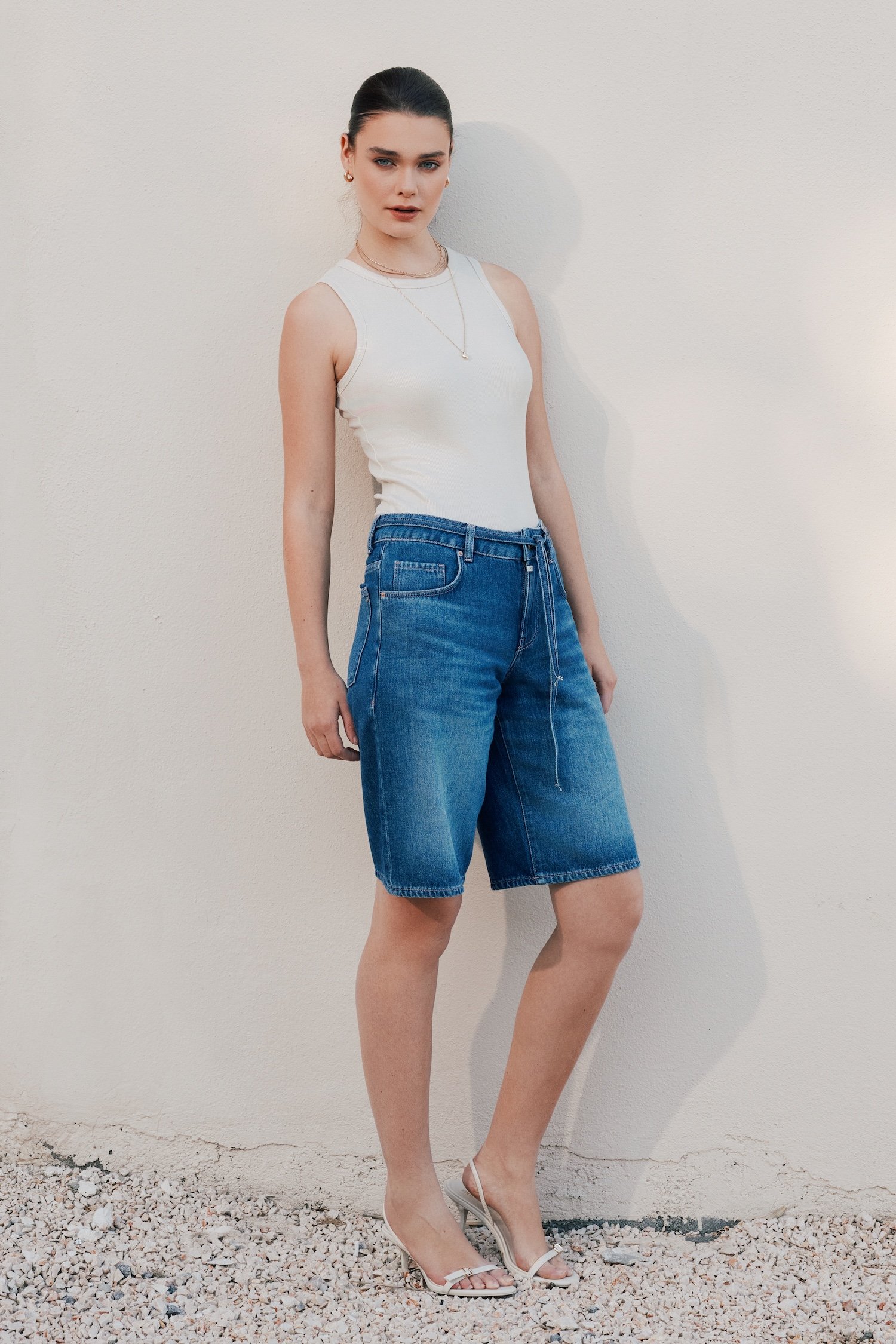 A woman in a white sleeveless top and blue denim shorts stands against a plain light-colored wall with her left leg slightly bent. She is wearing high-heeled sandals and has her dark hair pulled back. She also has layered necklaces and earrings, and is looking directly at the camera.