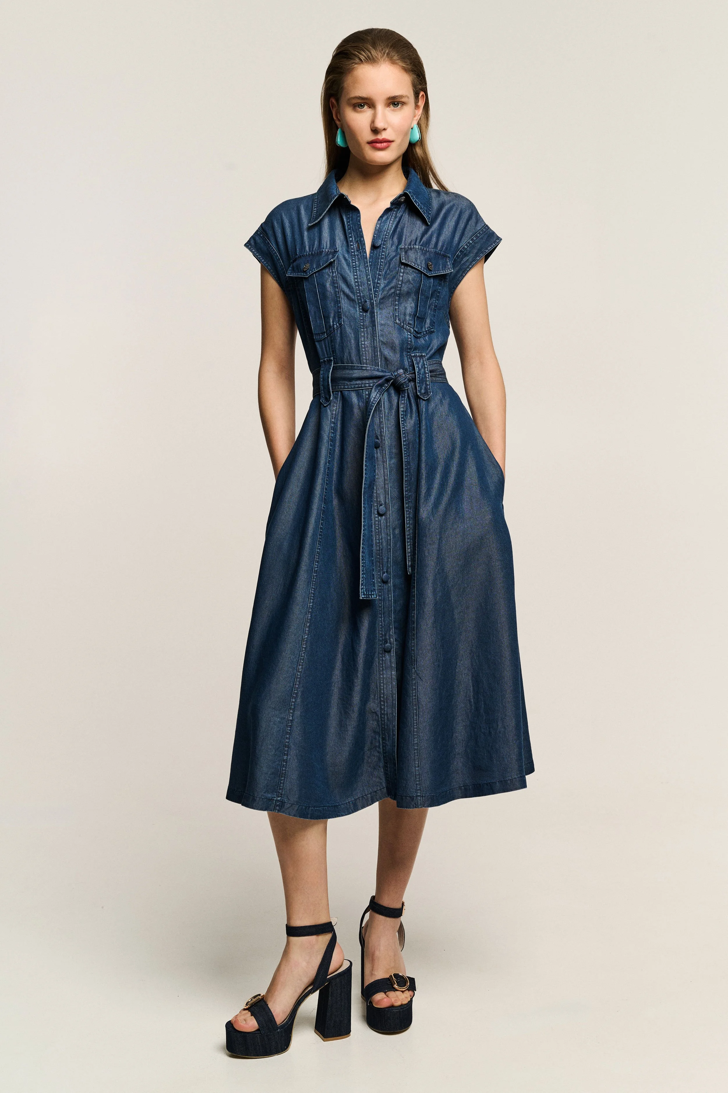 A woman standing against a plain light background, wearing a sleeveless denim dress with a collar, pockets on the chest, a waist tie, and a long skirt, paired with black high-heeled sandals with ankle straps.