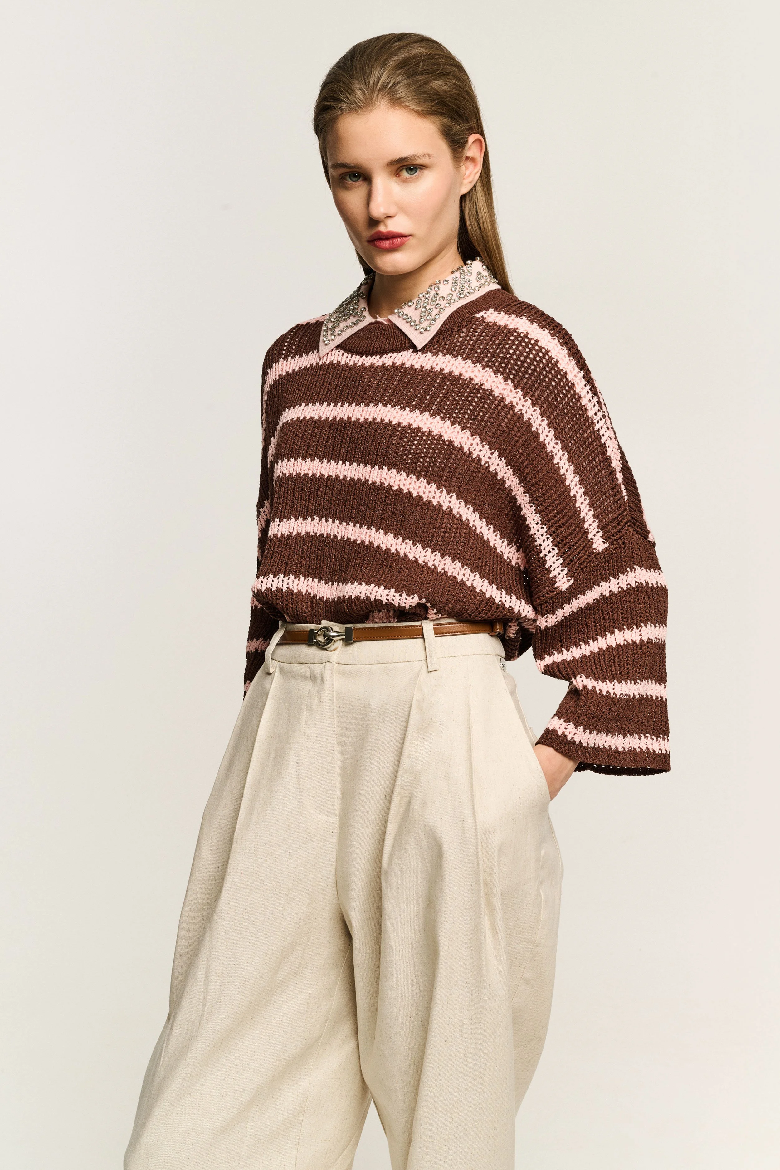 A young woman with blonde hair wearing a brown and pink striped sweater with a bejeweled collar, beige high-waisted wide-leg pants, and an elegant belt, standing against a plain light background.