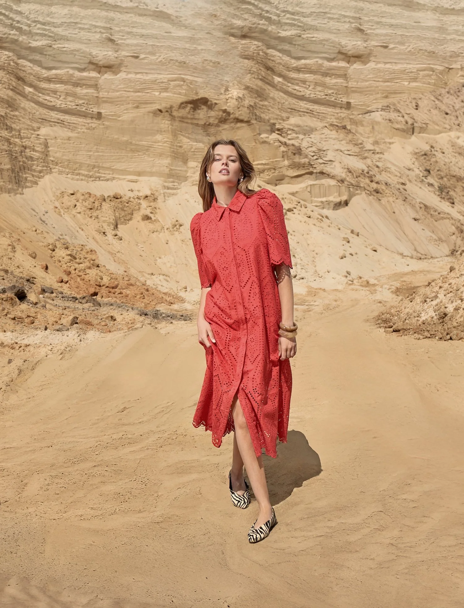 A woman in a red lace dress and zebra-print high heels standing in a desert landscape with large sand dunes and rock formations in the background.