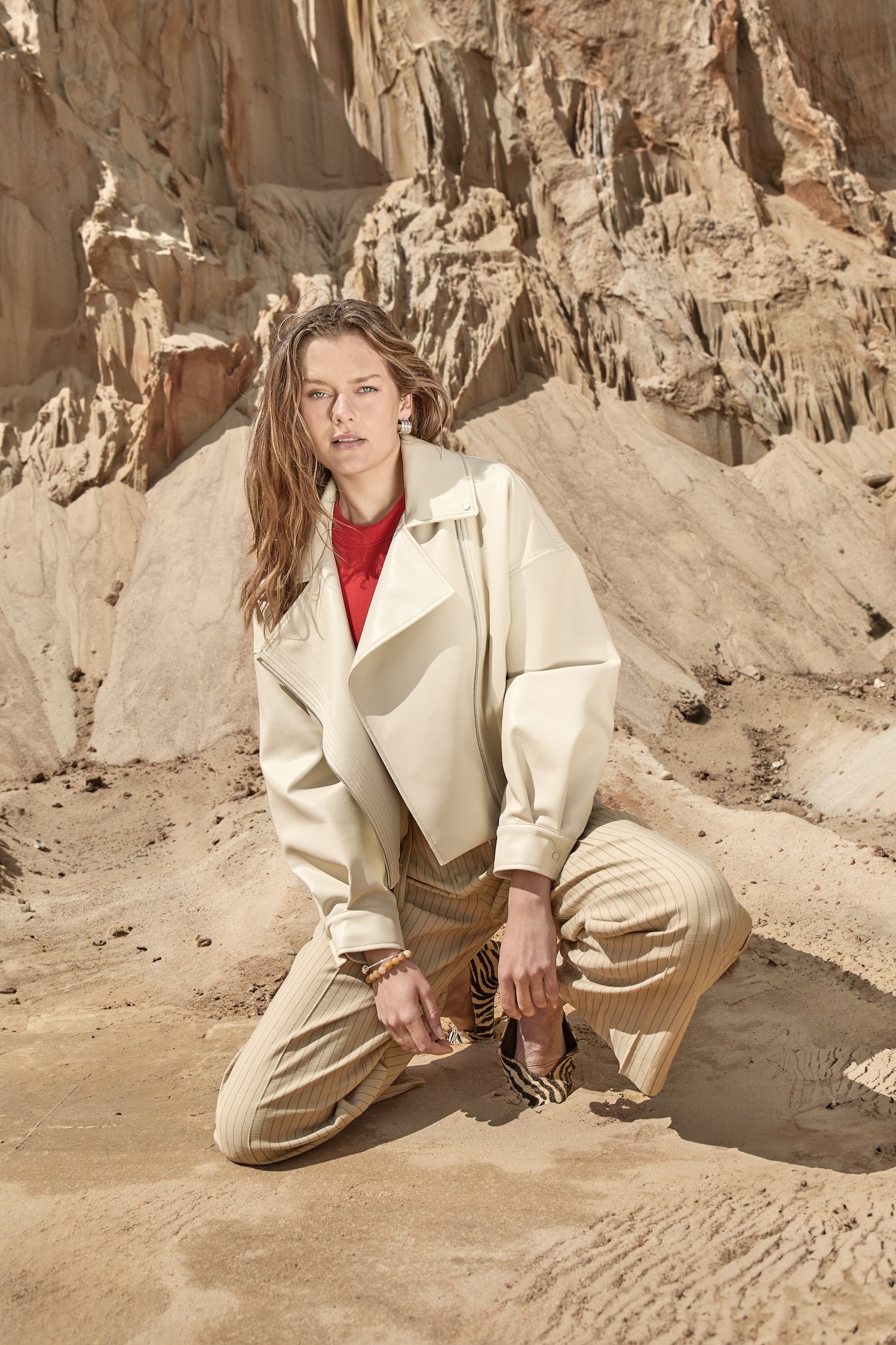 A woman with long wavy hair wearing a cream leather jacket, red top, and beige pinstripe pants kneeling on sandy terrain in a desert with rocky cliffs in the background.