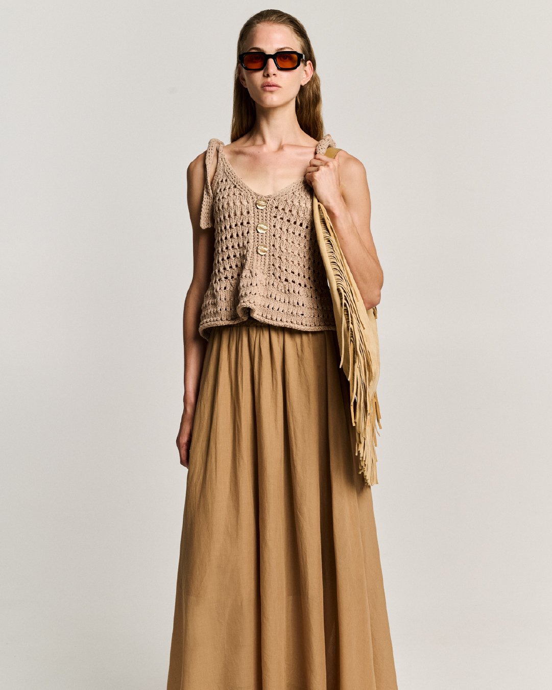 A woman wearing sunglasses, a beige crochet sleeveless top with buttons, a beige fringe bag, and tan wide-leg pants, standing against a neutral background.