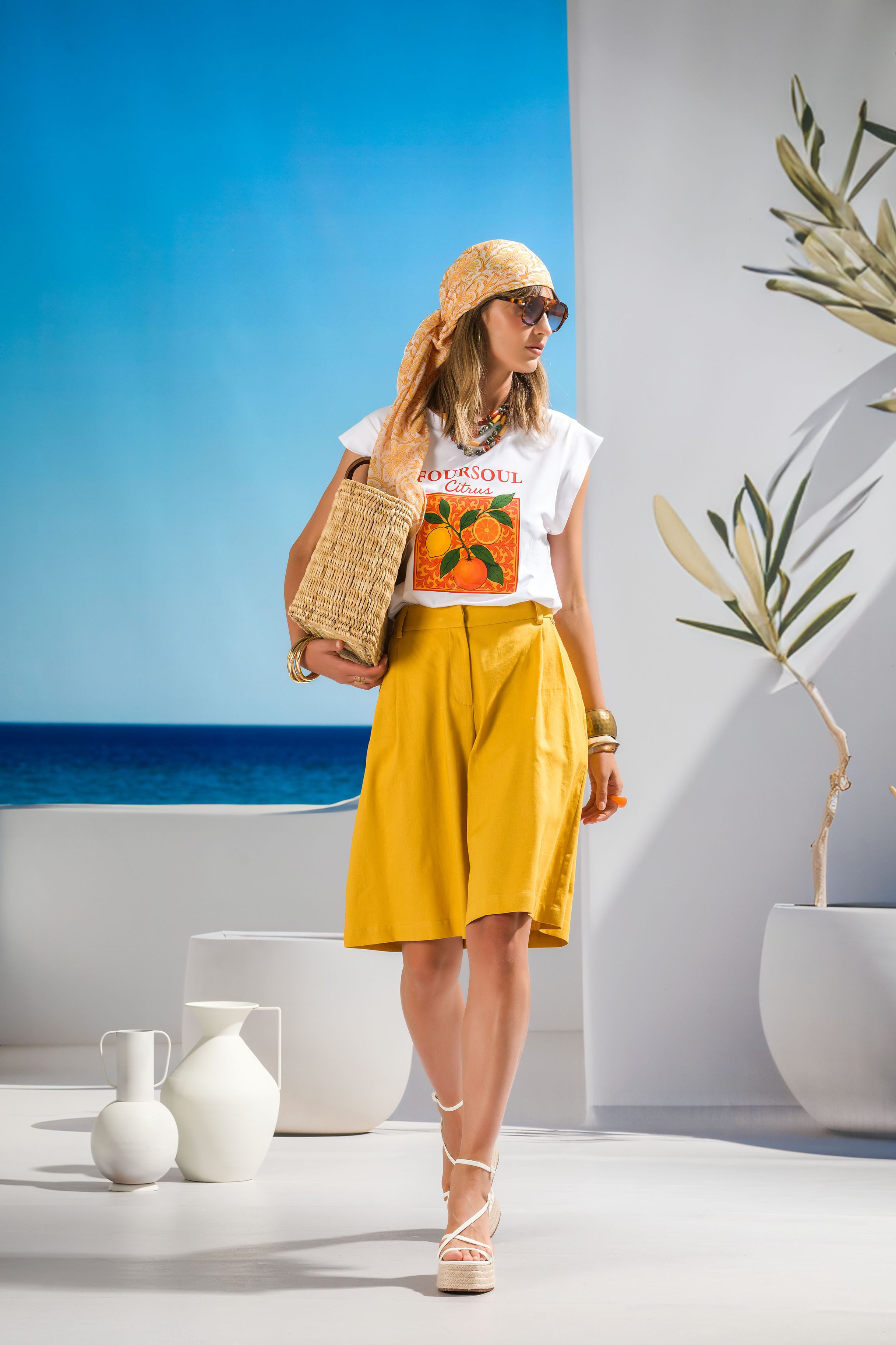 A woman in a white t-shirt with a citrus graphic, yellow high-waisted skirt, and white platform shoes stands in a bright, minimalist setting with white and blue backgrounds and white vases. She accessorizes with sunglasses, a patterned headscarf, colorful jewelry, and a woven tote bag, with some greenery visible on the right side of the image.