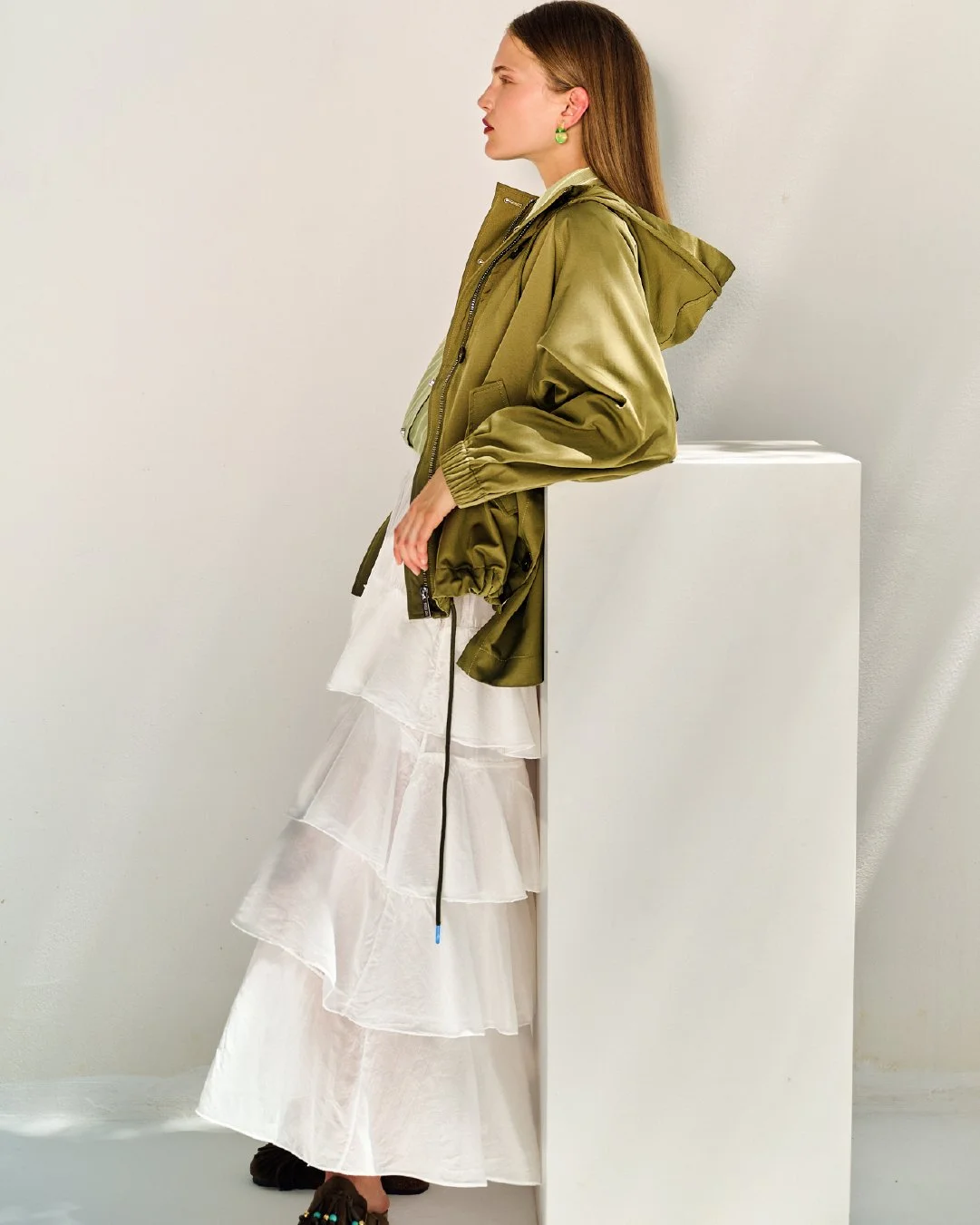 Woman standing in profile, wearing an olive green jacket and a tiered white maxi skirt, leaning against a white pedestal in a minimalist setting.
