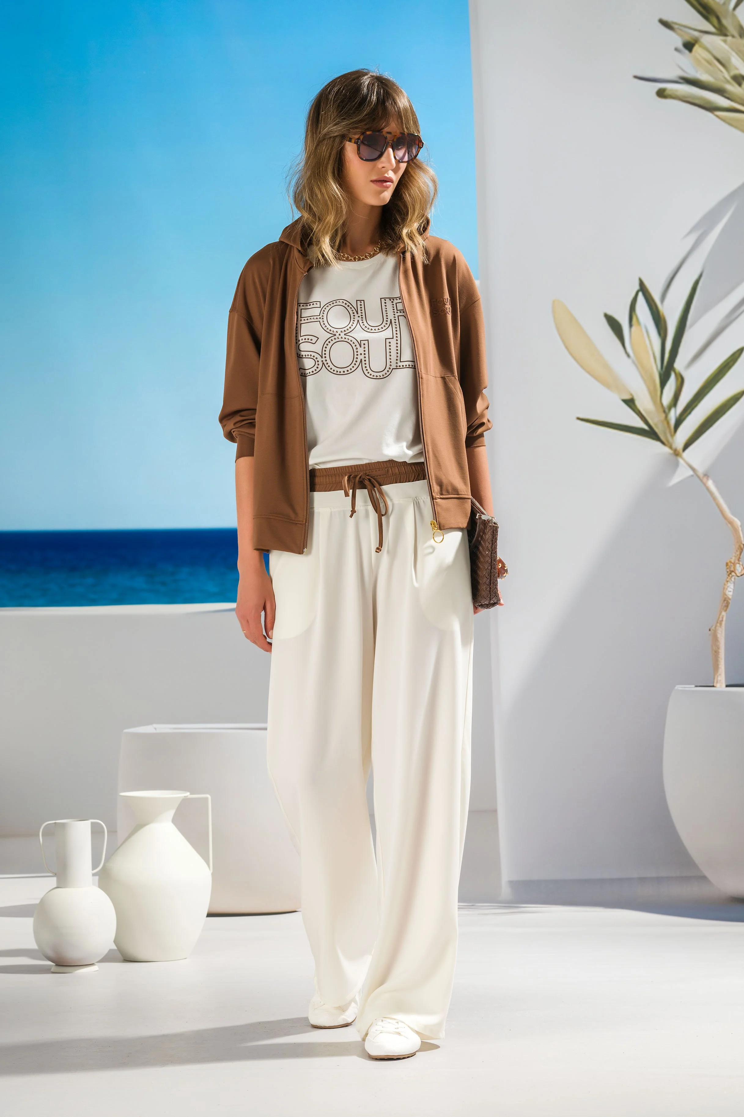A woman with wavy brown hair wearing large sunglasses, a brown jacket over a white t-shirt with a dotted print, wide-leg white pants, and white sneakers, standing indoors in front of a backdrop with a blue sky and ocean scene, alongside white decorative vases and a potted plant.