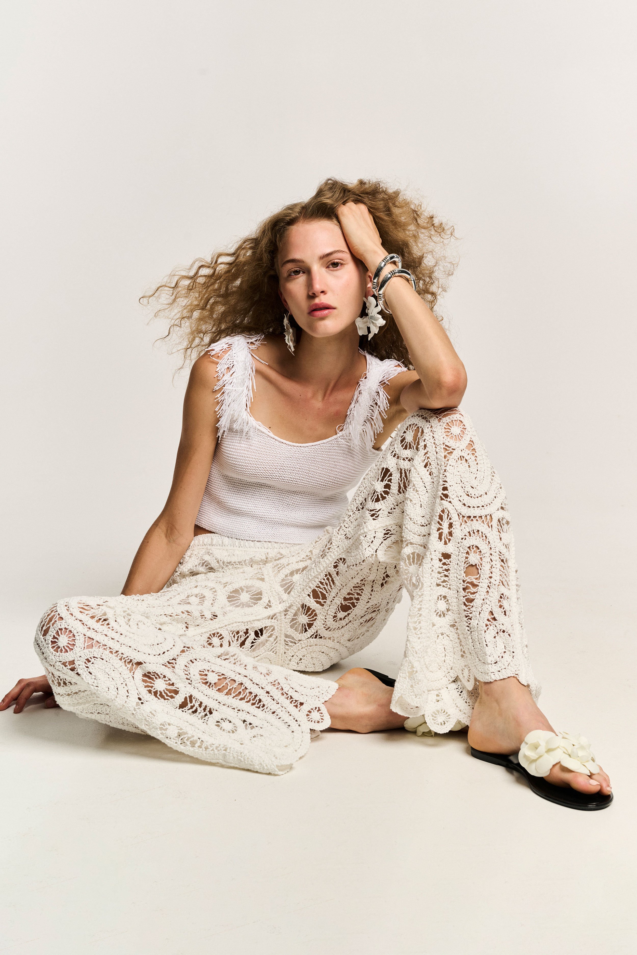 A woman sitting on the floor against a plain white background, wearing a white fringed top and lace patterned pants, with her left hand resting on her head and wearing large earrings and chunky bracelets, with a floral embellishment on her footwear.