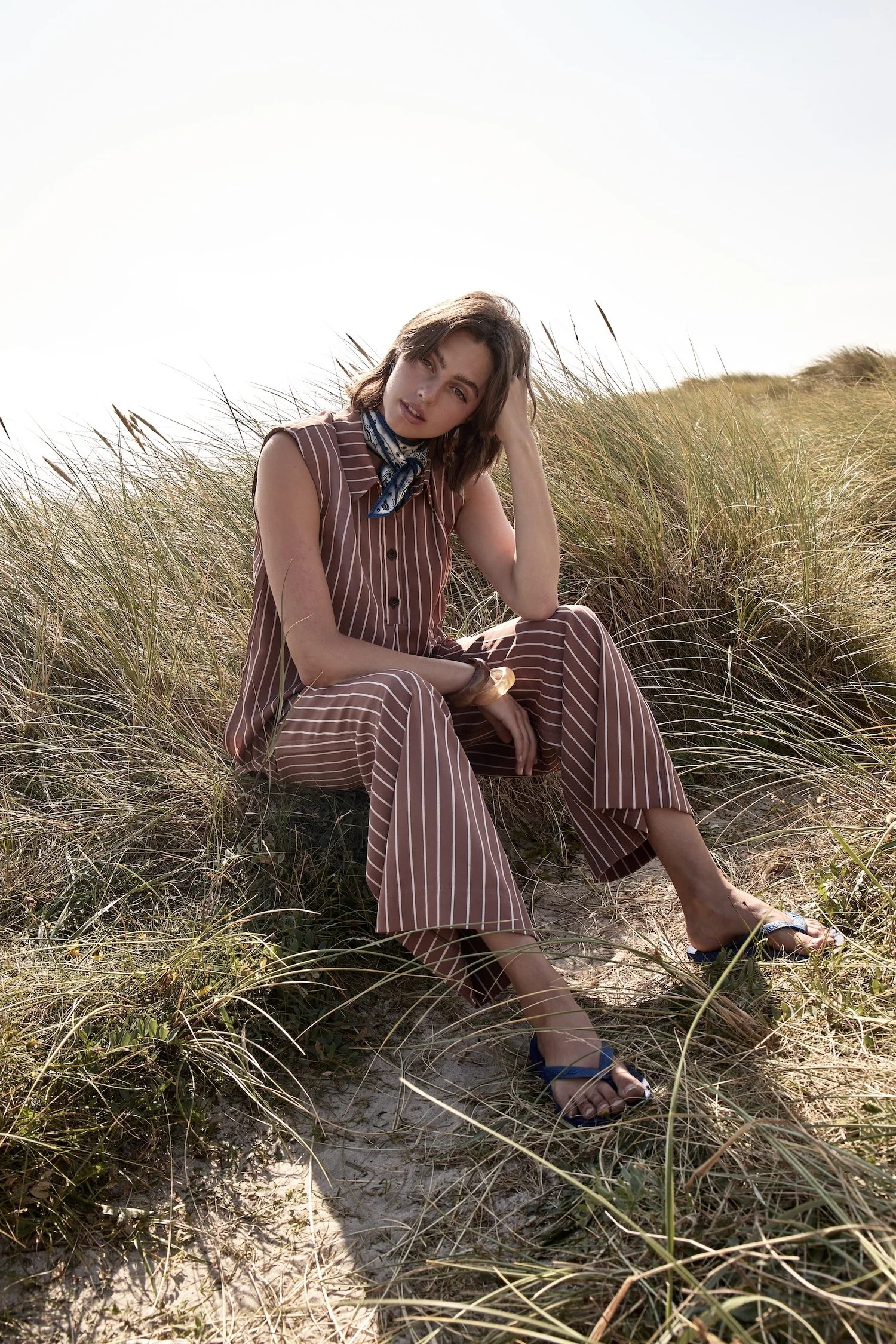 A young woman with short brown hair sits on a sandy and grassy area outdoors, wearing a brown and white striped sleeveless jumpsuit, a blue patterned scarf around her neck, and sandals. She is resting her head on her left hand and looking at the camera with a casual expression.