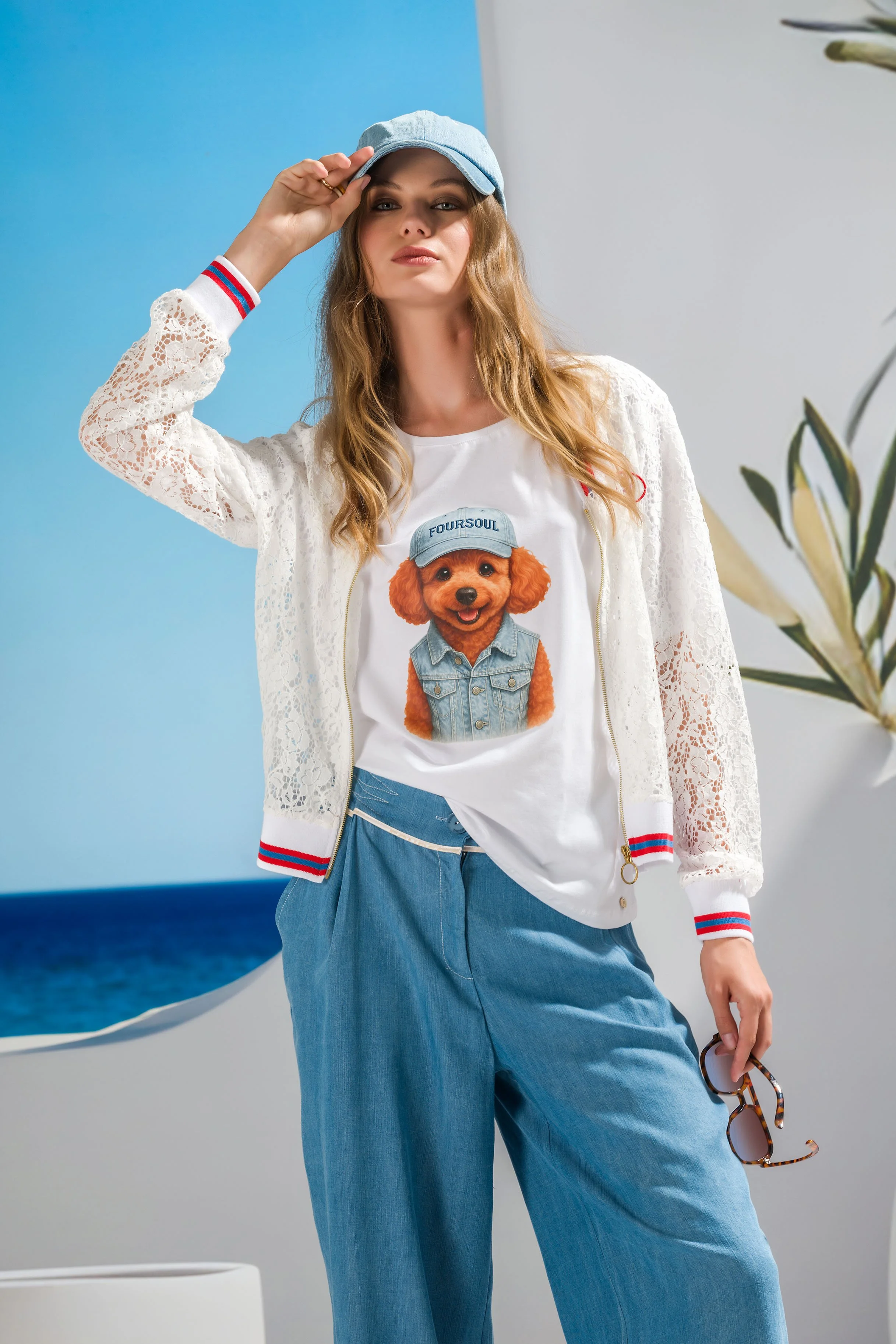 Young woman with wavy red hair wearing a denim cap, white lace jacket, graphic t-shirt with a dog in a denim vest, and loose blue pants, holding tortoiseshell sunglasses. She's standing indoors with a blue wall, white wall, plant, and abstract art in the background.