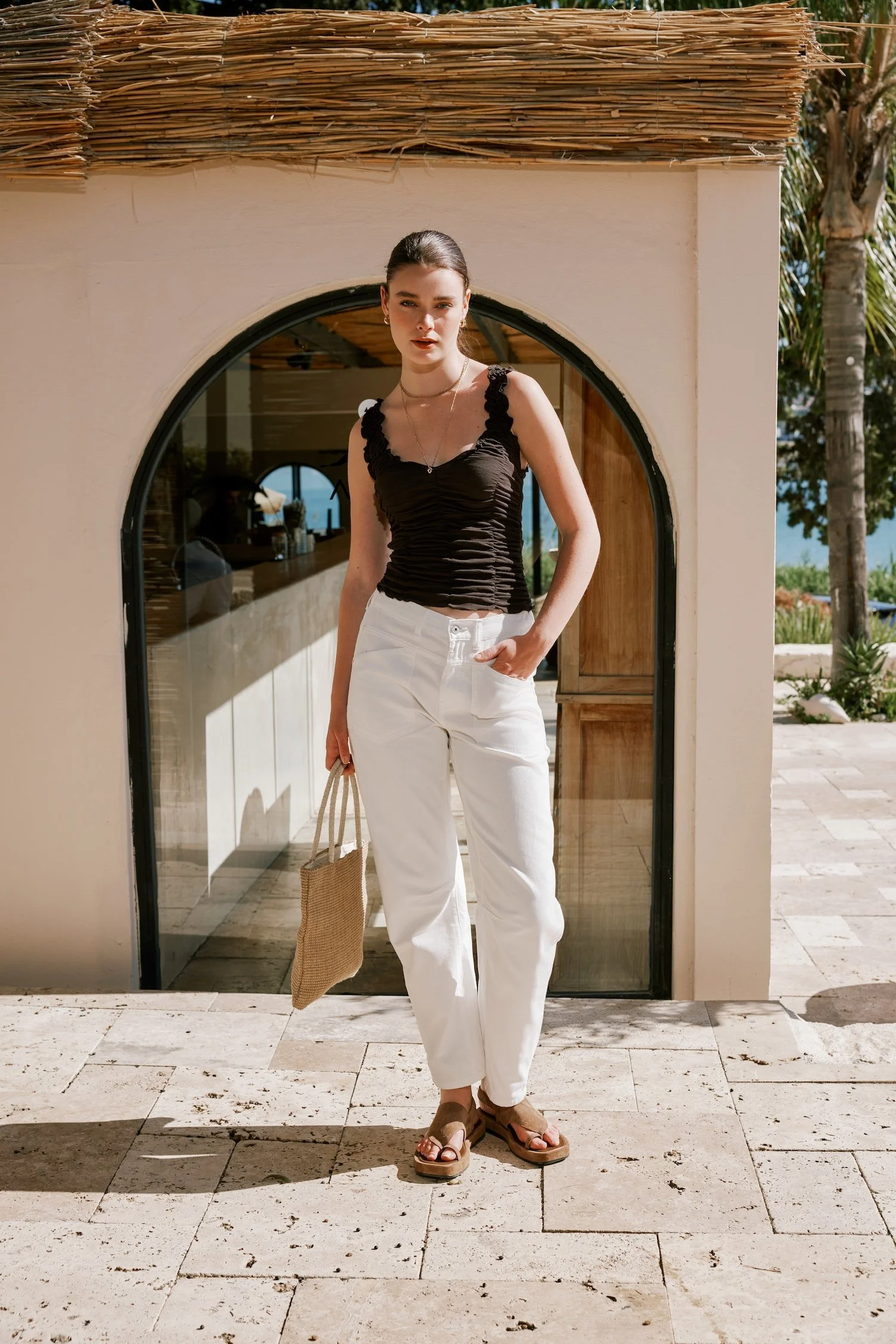 Young woman standing outside, wearing a black sleeveless top, white pants, brown sandals, holding a beige tote bag, with a background of a building, palm trees, and a view of the ocean in the distance.