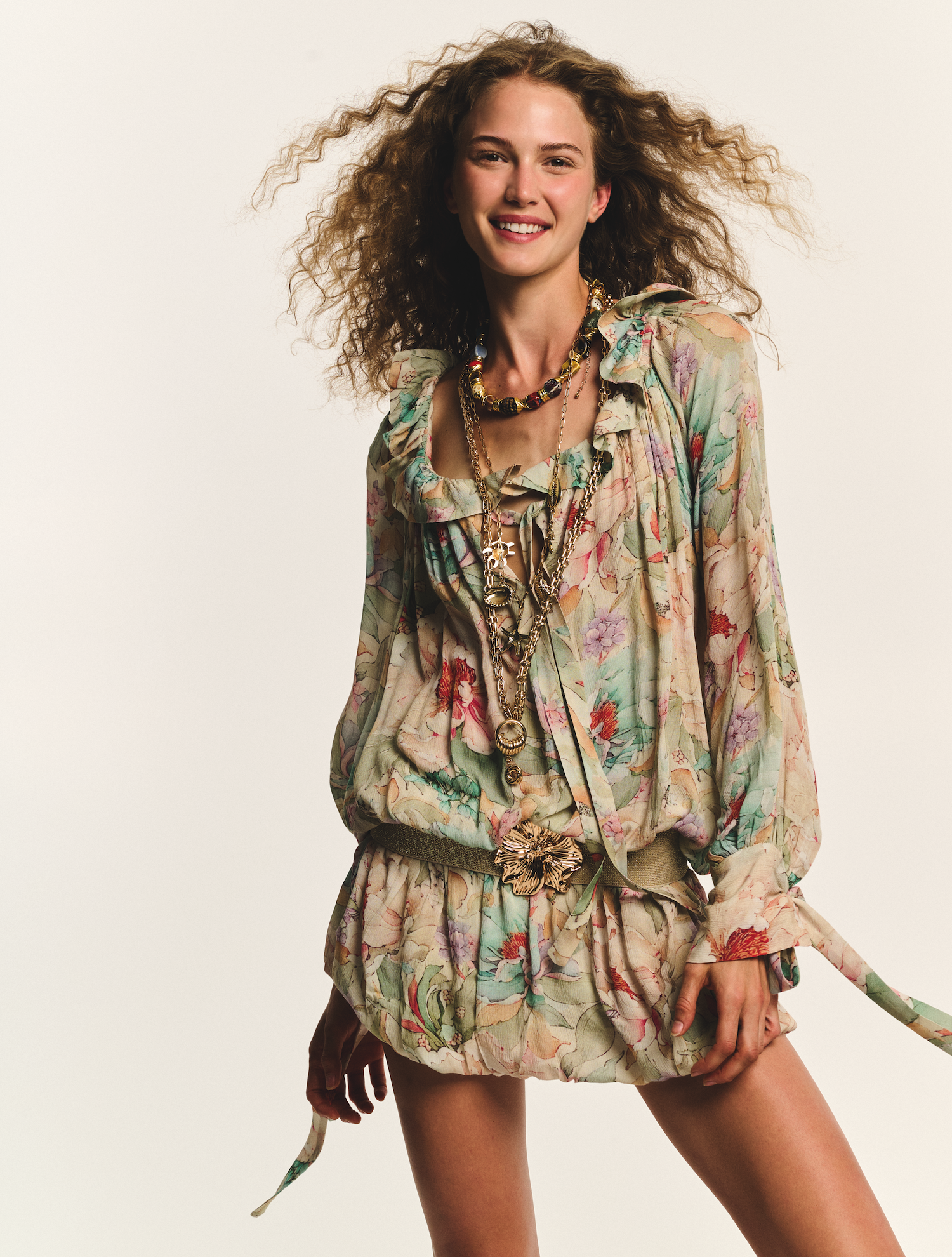 A young woman with curly hair smiling and wearing a floral dress with layered necklaces, holding a tied fabric in her hand.