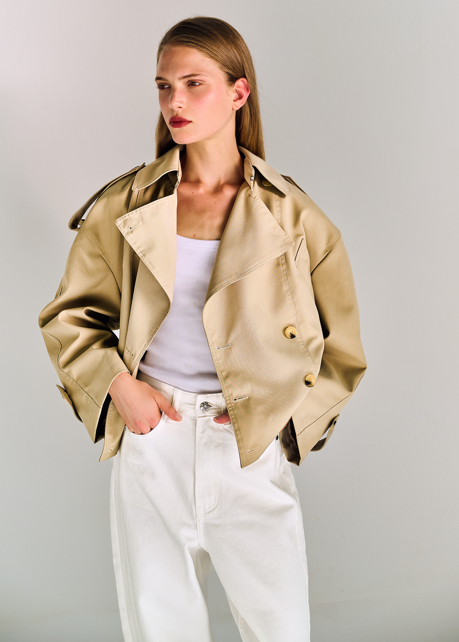 A young woman with long, straight, light brown hair wearing a beige trench coat, white top, and white jeans standing against a plain gray background.