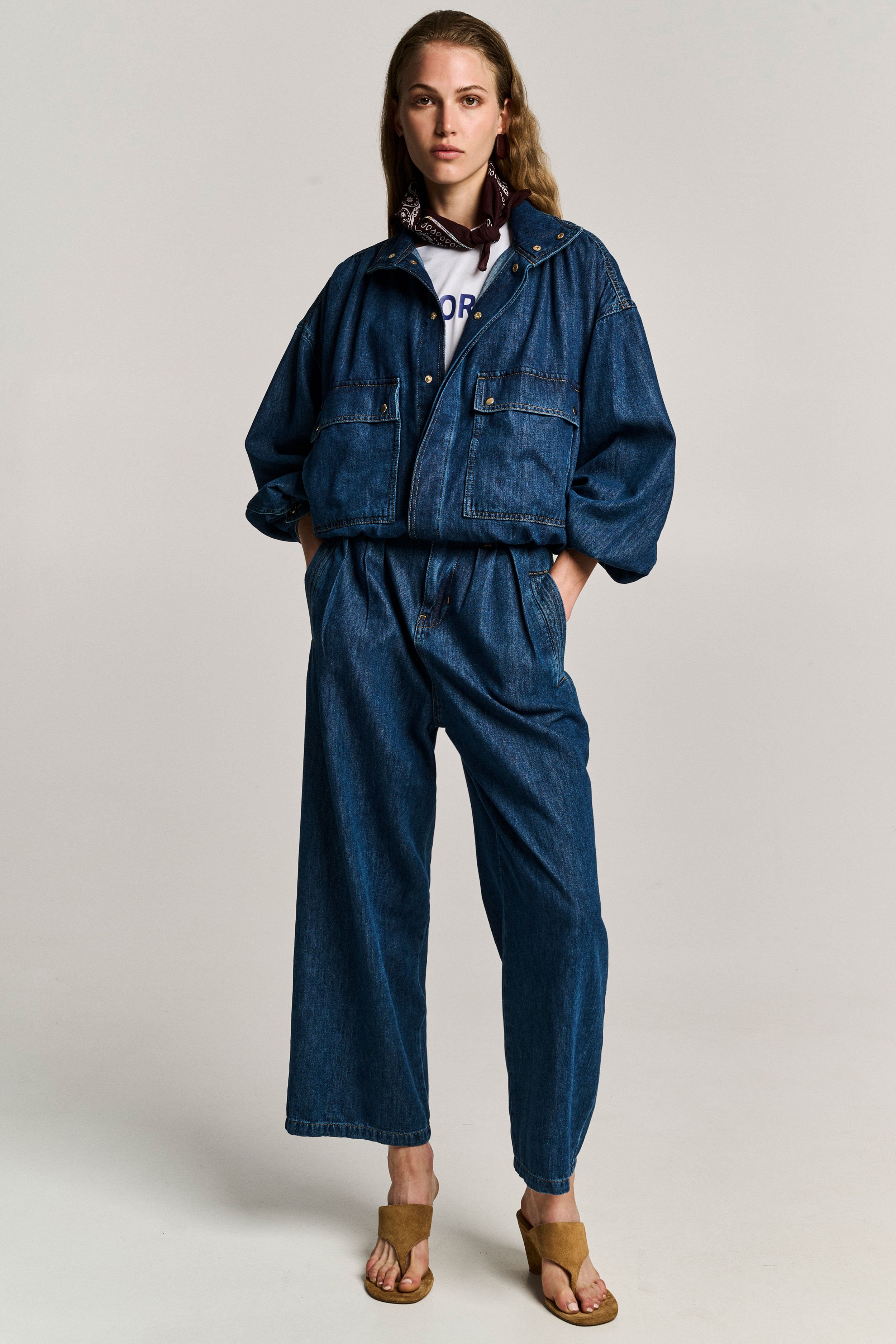 A full body shot of a woman wearing a denim jacket, matching denim pants, tan sandals, a white t-shirt, and a bandana around her neck, standing against a plain background.