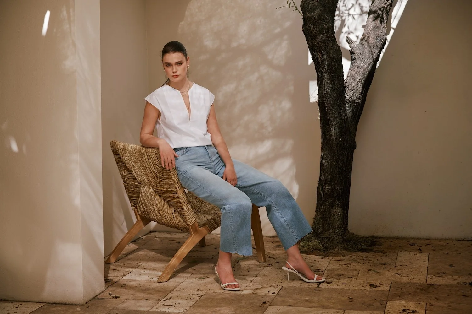 A woman sitting on a woven wooden chair in a minimalist setting with a tree and textured stone floor.