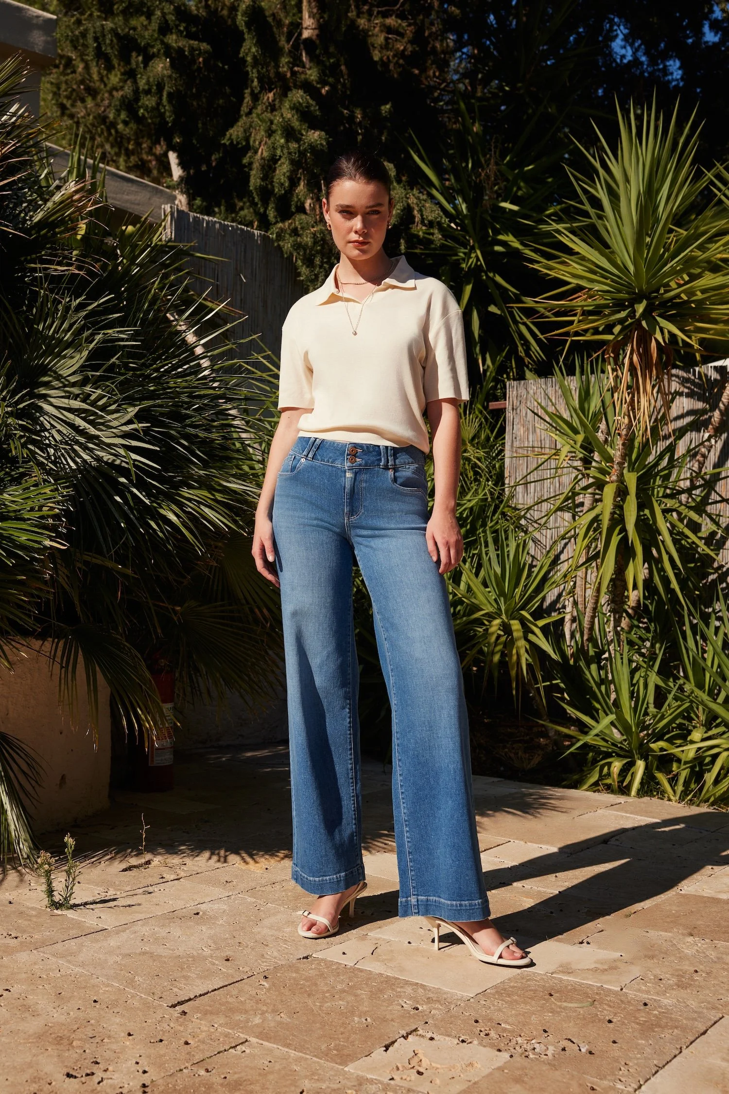 A woman standing outdoors surrounded by green tropical plants, wearing a cream-colored short-sleeve shirt, high-waisted blue jeans, and white open-toe high-heeled sandals.