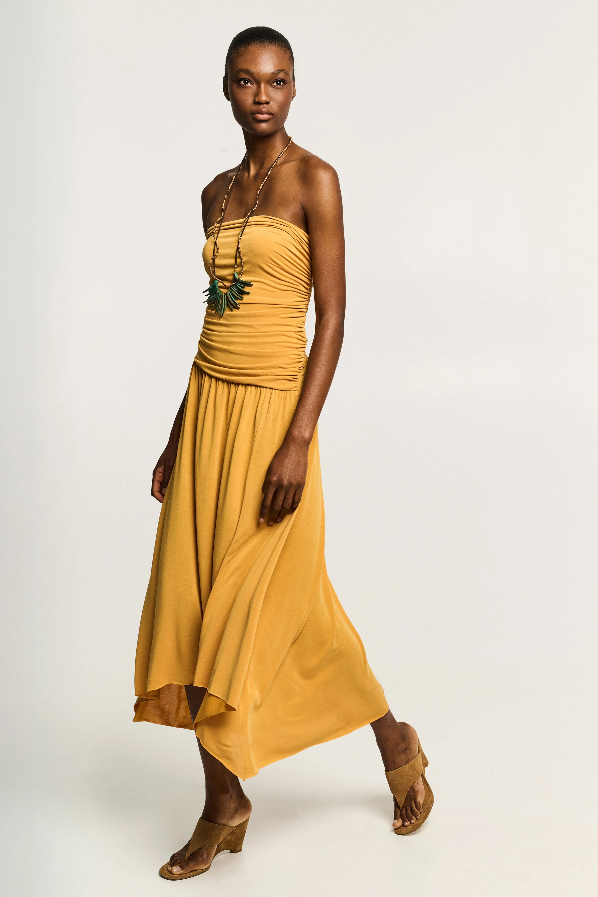 A woman with dark skin and short hair standing against a plain background, wearing a strapless, ruched yellow dress, accessorized with a long necklace and beige high-heeled sandals.