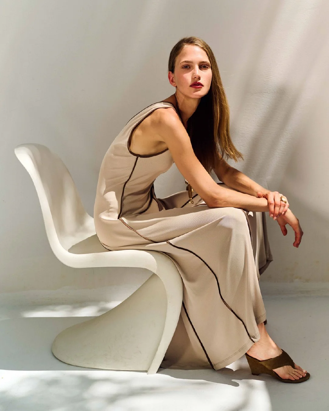 A woman sitting on a modern white chair, wearing a beige dress with black piping, with long brown hair, against a plain light-colored wall.