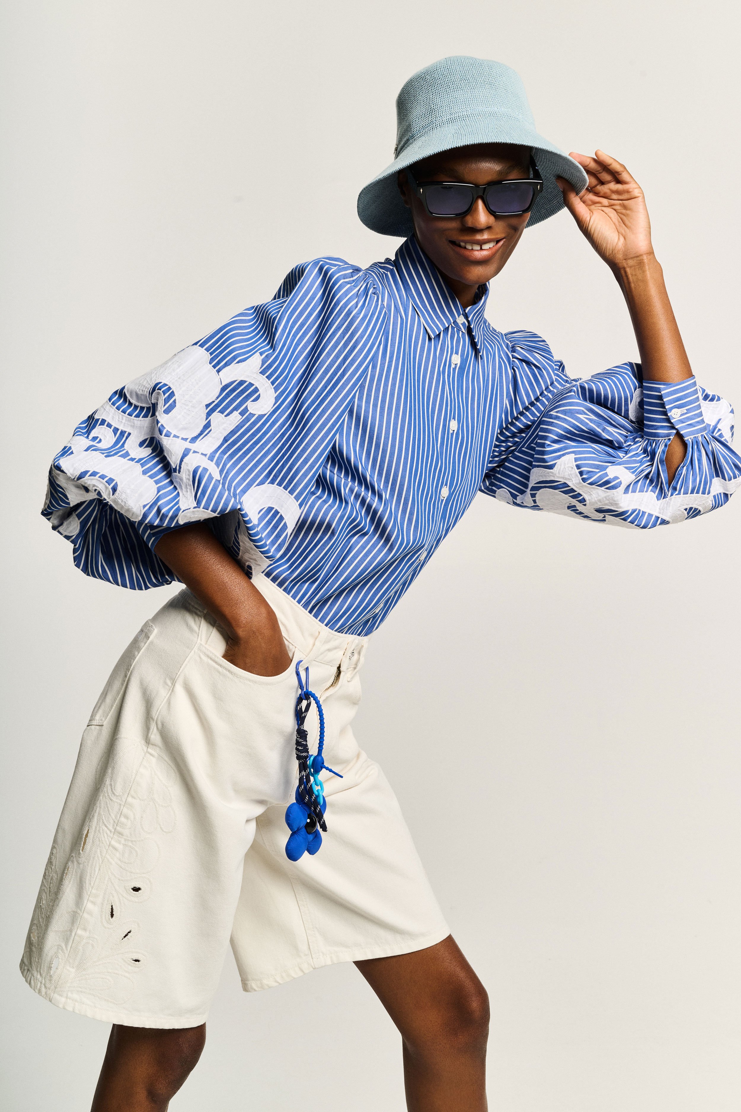 Person wearing a wide-brimmed hat, sunglasses, and a blue striped shirt with white details, posing with one hand on the hat and one leg slightly bent, against a plain light background.