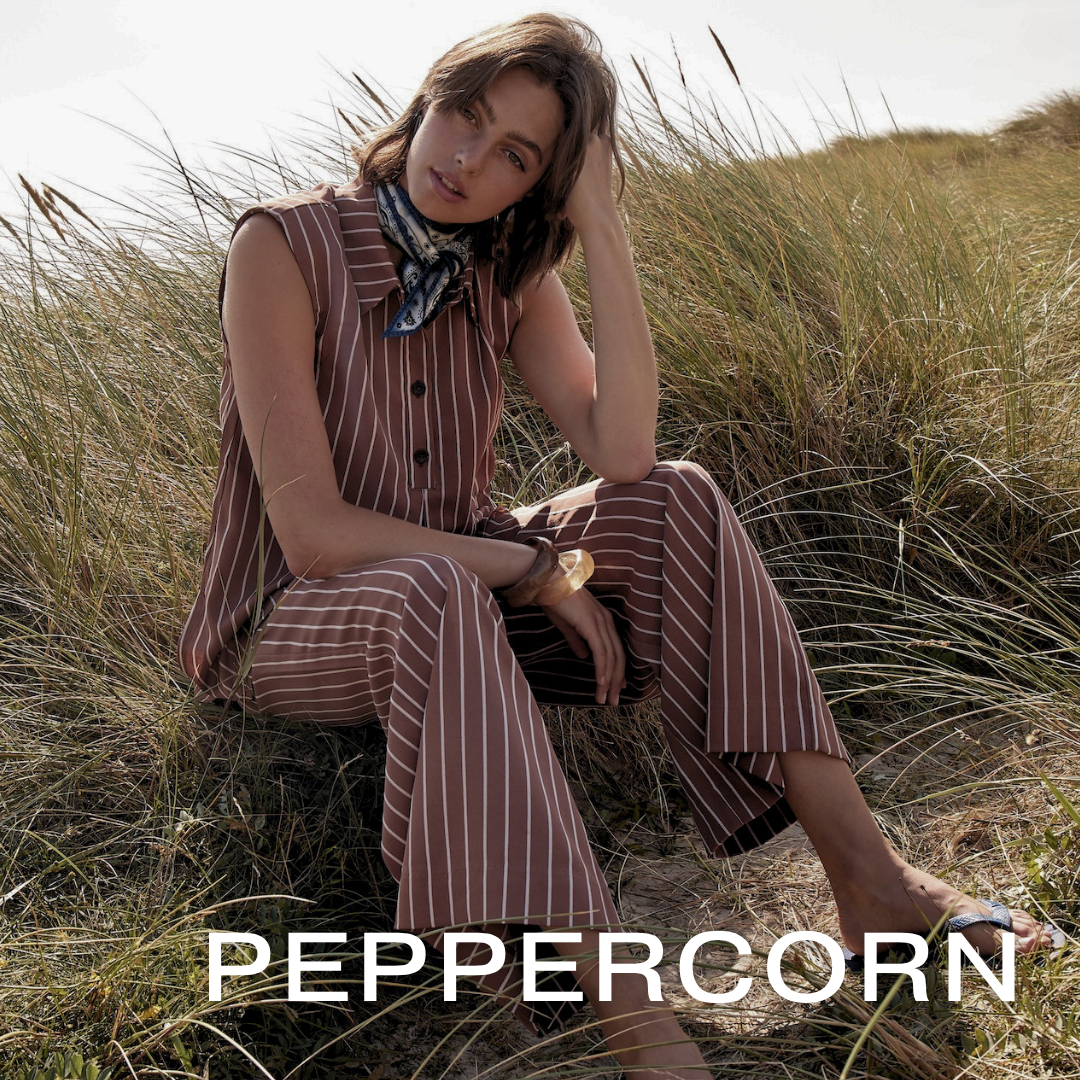 A young woman with short brown hair sitting on sandy ground surrounded by tall grass in a field, wearing a pinstriped brown jumpsuit, a blue patterned scarf, and sandals, with one hand resting on her head, looking at the camera.