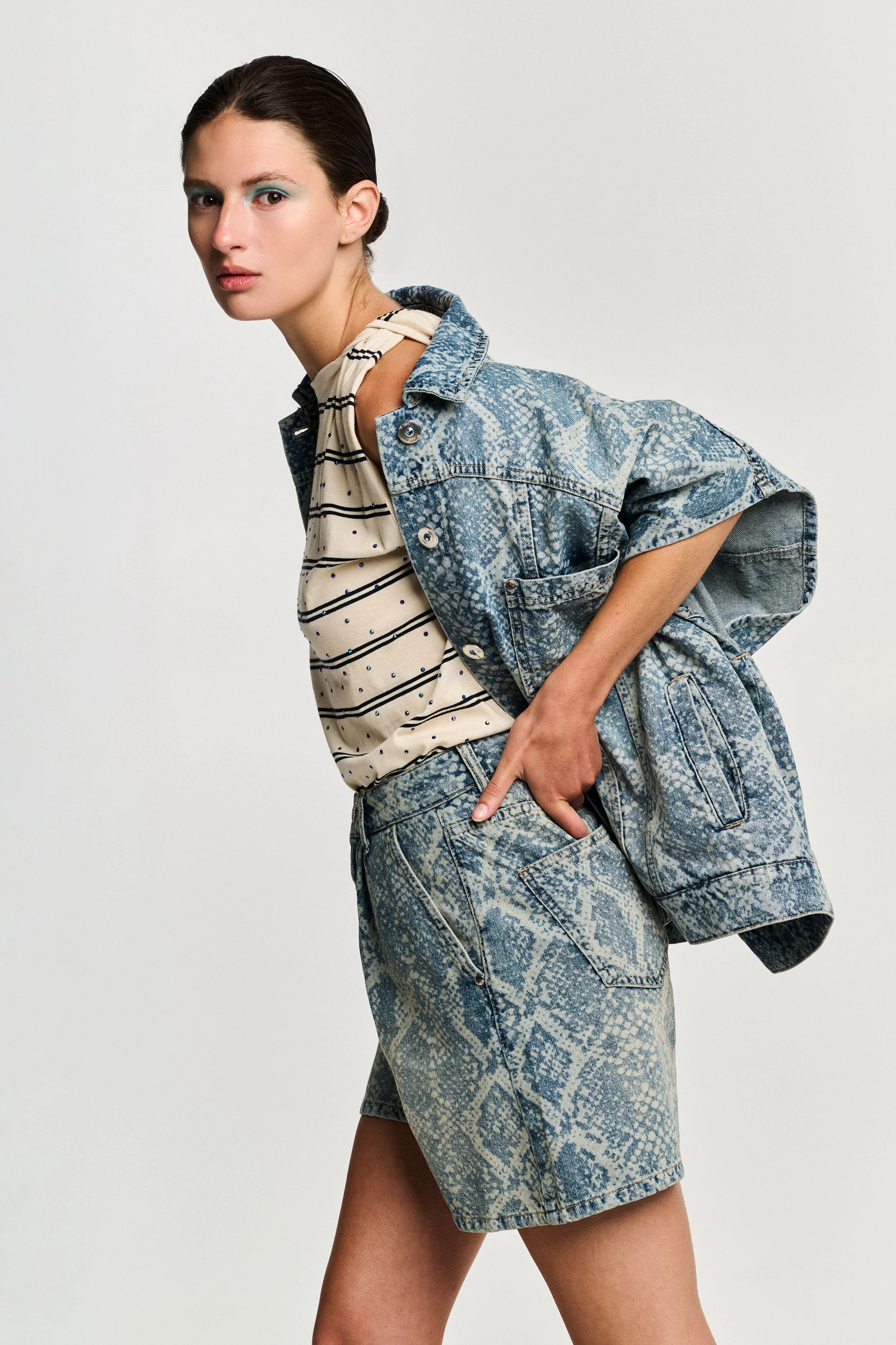 A young woman in denim jacket and shorts poses against a plain white background.