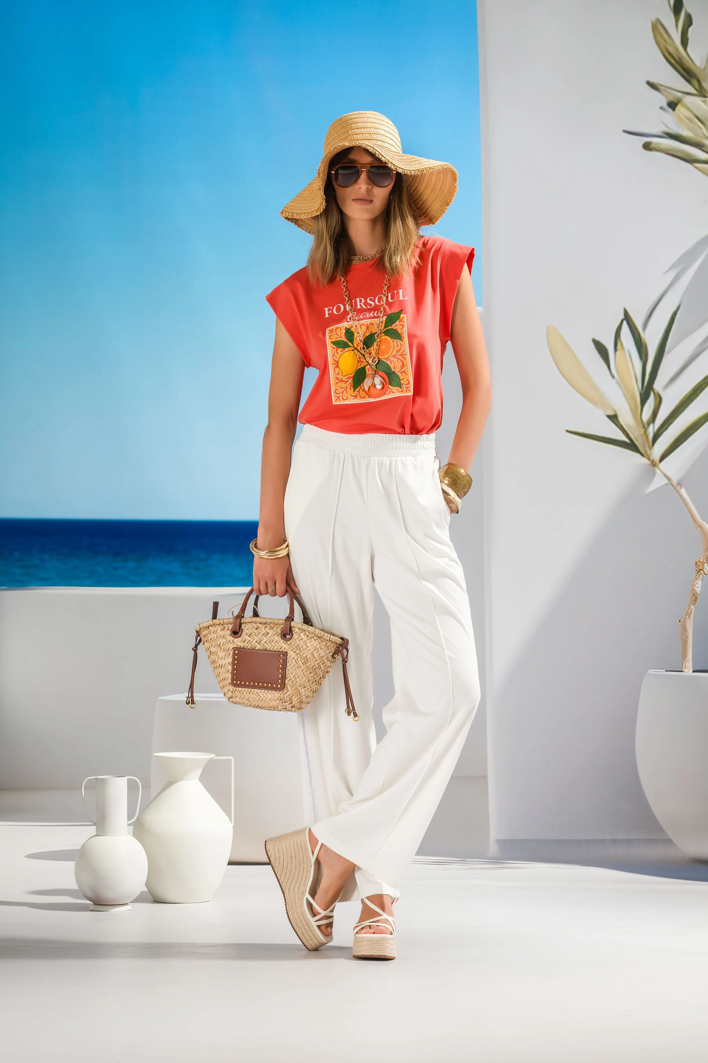 Woman wearing a straw hat, dark sunglasses, a red sleeveless top with a colorful fruit graphic, white high-waisted wide-leg pants, beige wedge sandals, gold jewelry, holding a woven straw bag, standing in a bright minimal setting with white vases and a plant, near a backdrop of the ocean.