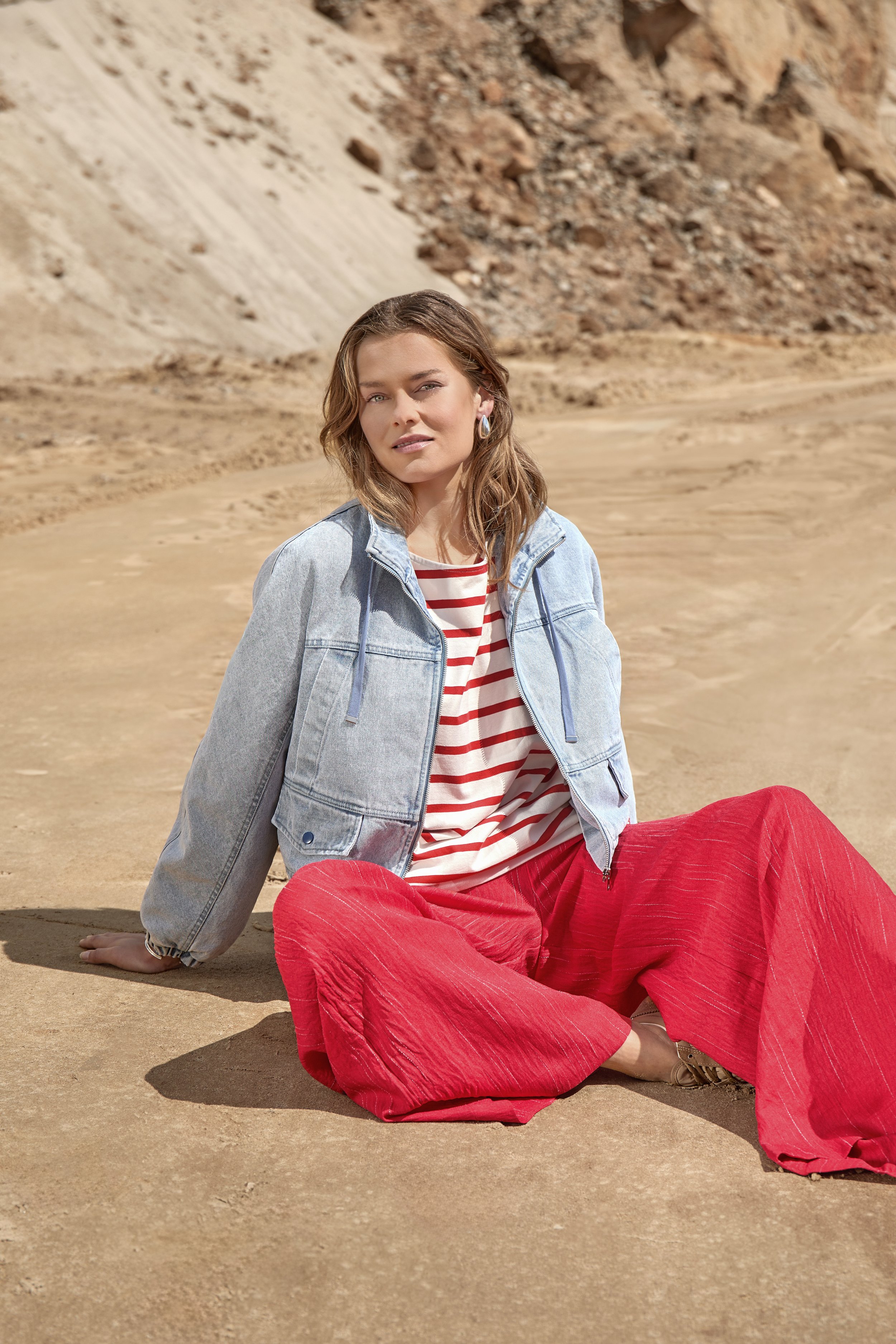A woman with wavy brown hair is sitting on the sandy ground in a desert landscape, wearing a denim jacket, a red and white striped shirt, and bright red pants. There are rocky hills or mountains in the background.