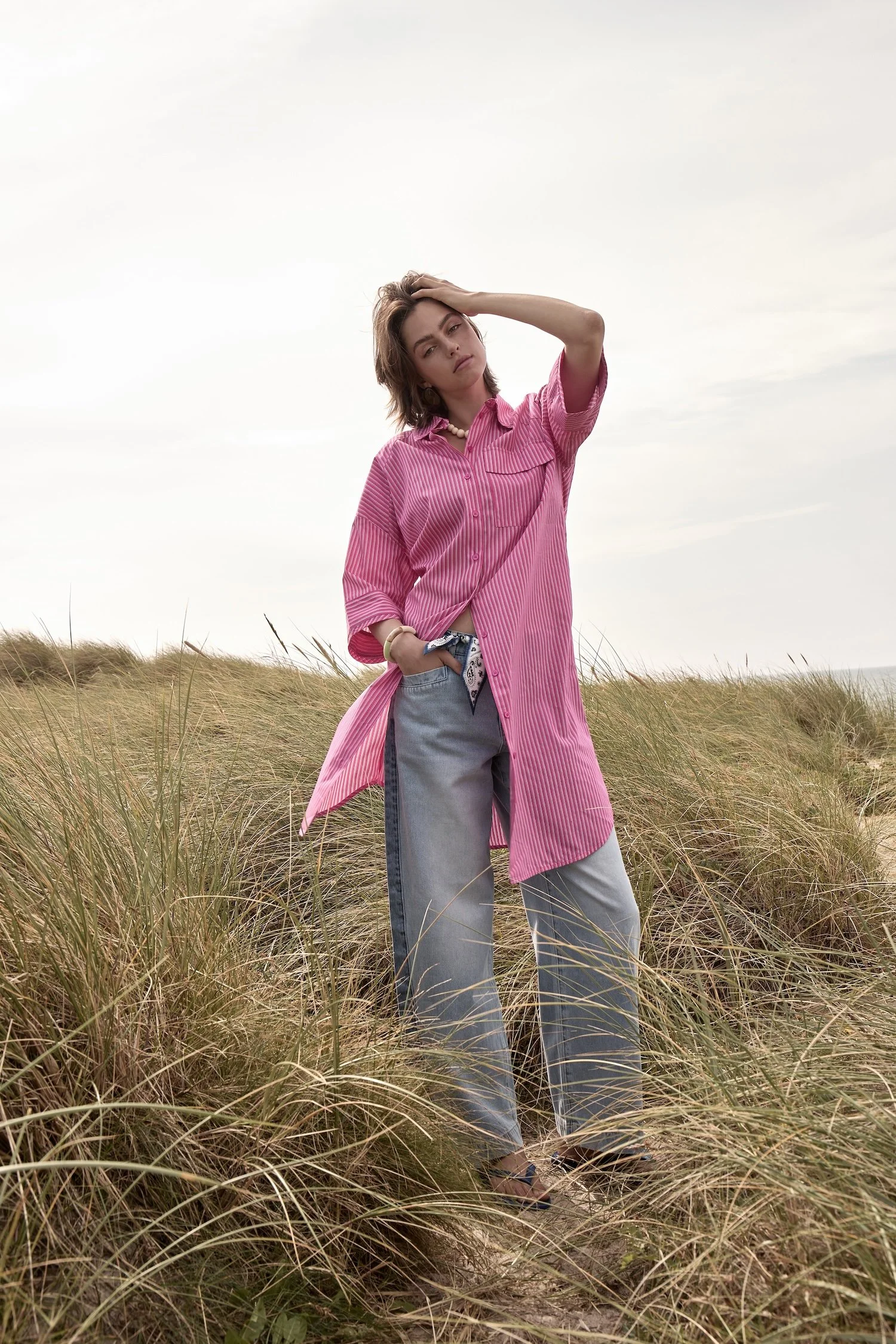 Young woman standing in a grassy field, wearing a pink striped oversized shirt and light jeans, with one hand on her head and looking at the camera.